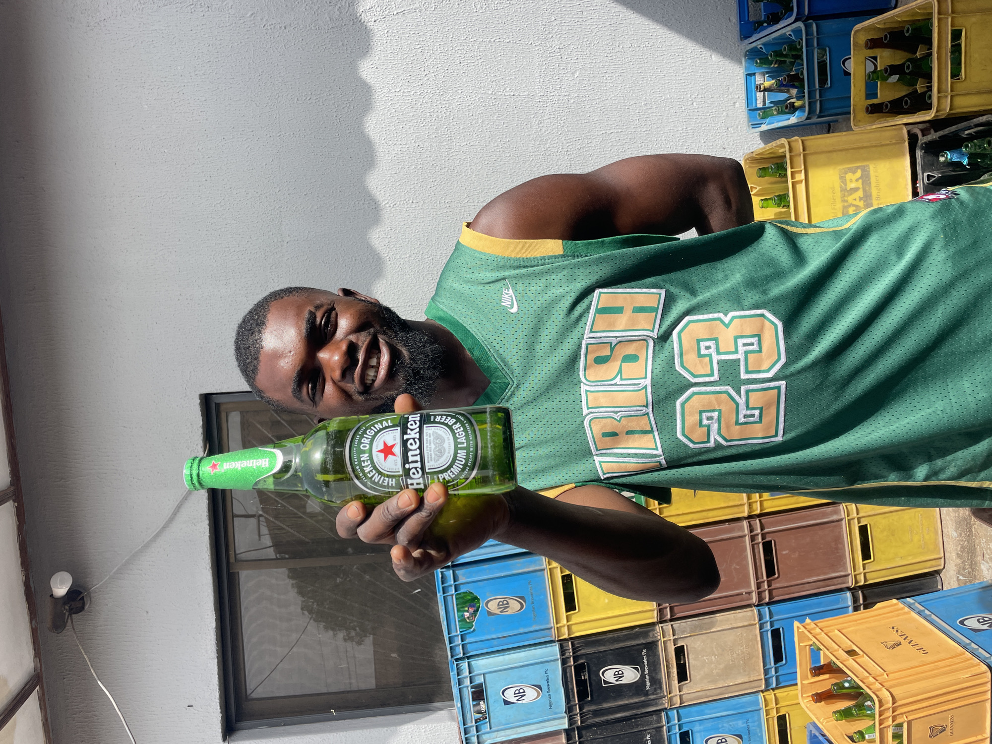 A medium shot shows a dark-skinned man holding a green Heineken beer bottle, positioned in front of rows of stacked beer crates. The man, wearing a green Irish jersey with the number 23, is smiling at the camera. Behind him is a white wall with a window and an outdoor light fixture. The beer crates, stacked high, display various logos, including "NB," "Guinness," and "WAR - Brighter life." Although the image location in Jos, Nigeria, is specified, there are no indicators of Jos or its specific features present in the image. The description is limited to what is directly visible.