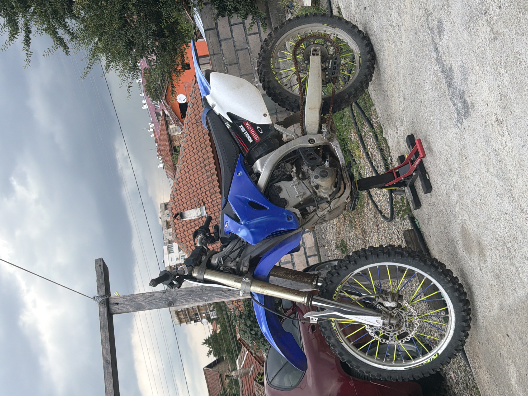 In an outdoor urban setting in Pavel Banya, Bulgaria, a blue and white dirt bike is parked on a concrete surface. The motorcycle is supported by a red and black stand. In the background, several residential buildings with red tile roofs are visible, along with trees and a cloudy sky. A portion of a maroon car is also visible to the left of the dirt bike. The scene appears to be a residential area.