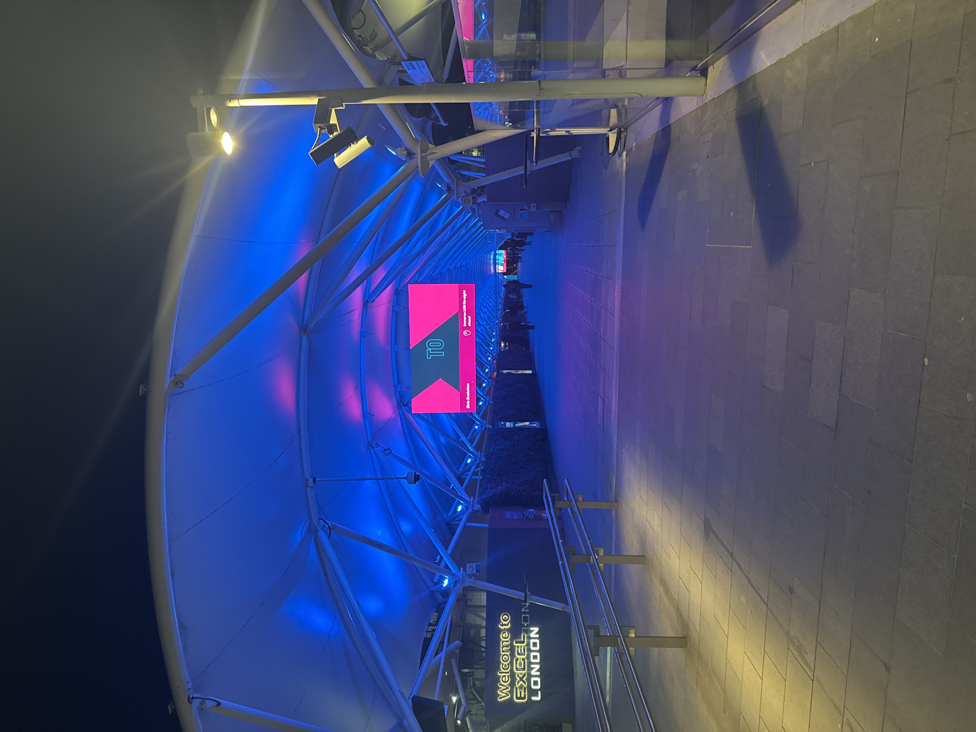 The image depicts the entrance to the ExCeL London exhibition and convention centre in London, United Kingdom, at night. The architecture is characterised by a tubular, canopy-like structure, illuminated with blue and pink lighting. The walkway is paved with grey tiles. A sign reads "Welcome to ExCeL LONDON". A digital display is present in the scene. People can be seen in the distance along the walkway. Street lights illuminate the area, casting shadows on the pavement. The sky is dark in the background.