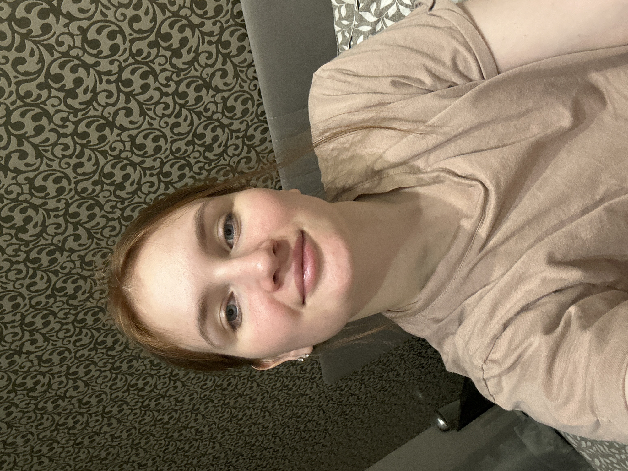 A young woman with reddish-brown hair and light skin is facing the camera. She has blue eyes and pink lips. She's wearing a light brown T-shirt. The background consists of a wall with a dark brown patterned wallpaper and a grey headboard of a bed. The location is Chaikovsky, Russia.