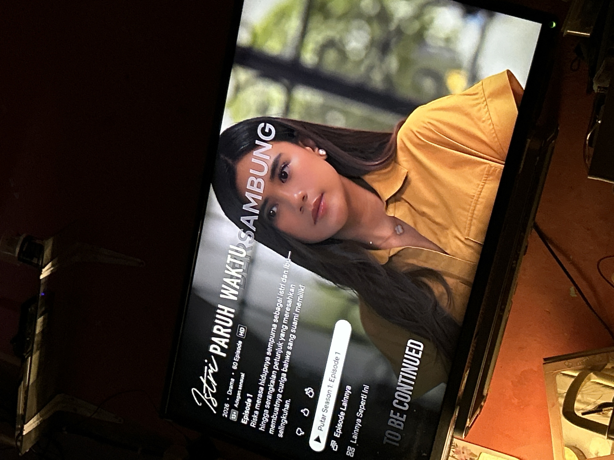 The image shows a close-up of a television screen displaying a scene from the Indonesian drama series "Istri Paruh Waktu Sambung." The screen is positioned at an angle, suggesting it is in a dimly lit room, possibly a living room. The show's title is prominently displayed in a stylized font, alongside descriptive text about the episode, including "Episode 1" and a brief synopsis. A young woman with long, dark hair is the main subject on screen, wearing a yellow collared shirt. Her gaze is directed away from the viewer. Below the episode information, there are interface elements, including a play button labeled "Putar Season 1, Episode 1" and options for "Episode Lainnya" (Other Episodes) and "Lainnya Seperti Ini" (More Like This). The bottom of the screen displays "TO BE CONTINUED," indicating the end of an episode. The television itself rests on a dark surface, and the surrounding environment appears to be a home interior. No specific landmarks or locations within Depok, Indonesia, are identifiable from the image.