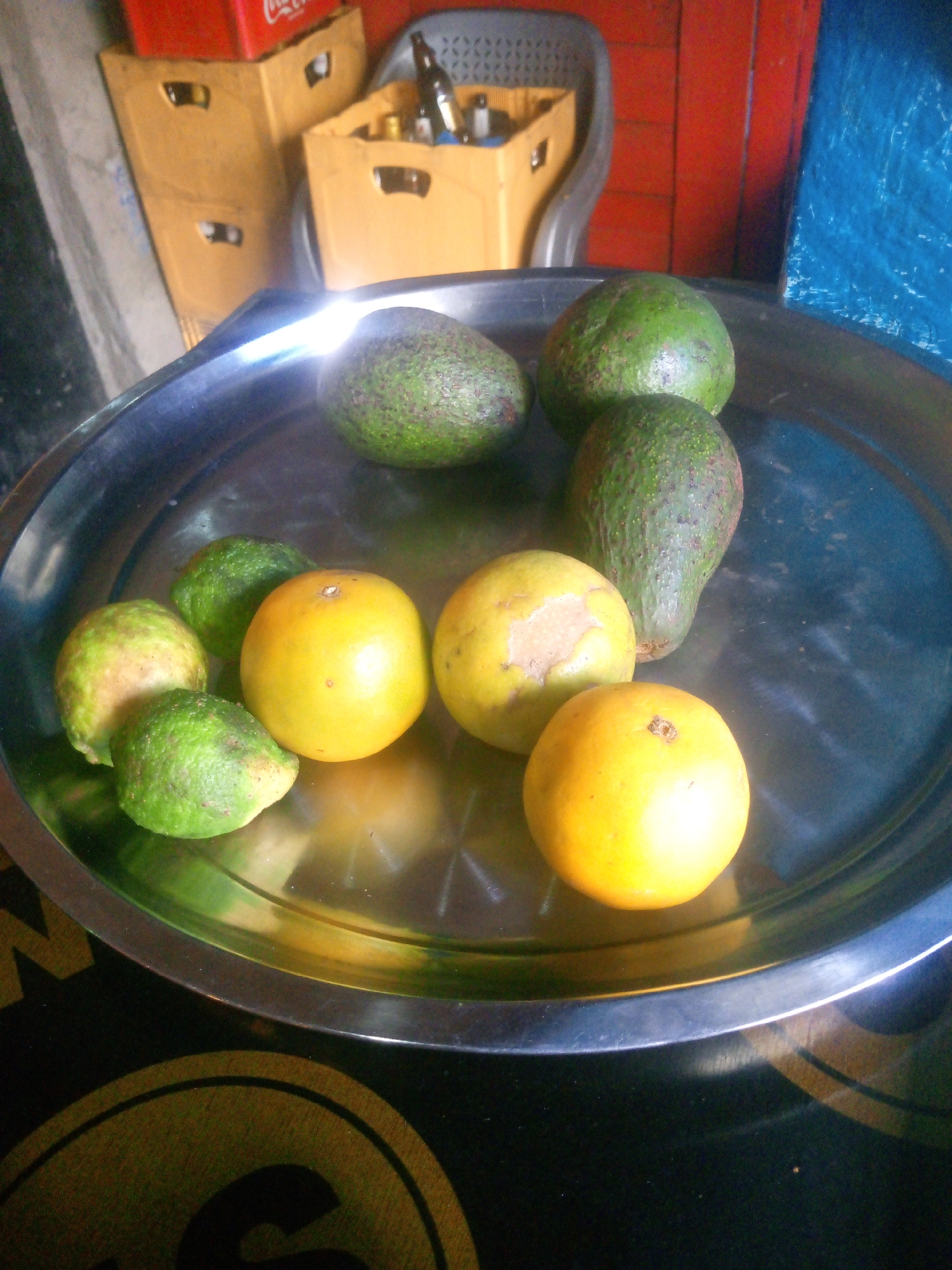 A high-angle shot shows a silver metal tray laden with produce: four avocados of varying sizes and ripeness, and three yellow oranges. The avocados are green and textured, while the oranges are smooth and bright yellow. The tray is placed on a dark surface with a gold design visible on the tray's edge. In the background are crates stacked against a wall, with a Coca-Cola branded crate on top and other crates containing bottles. A gray plastic chair is visible. The lighting is diffused. Nairobi, Kenya is provided as location.