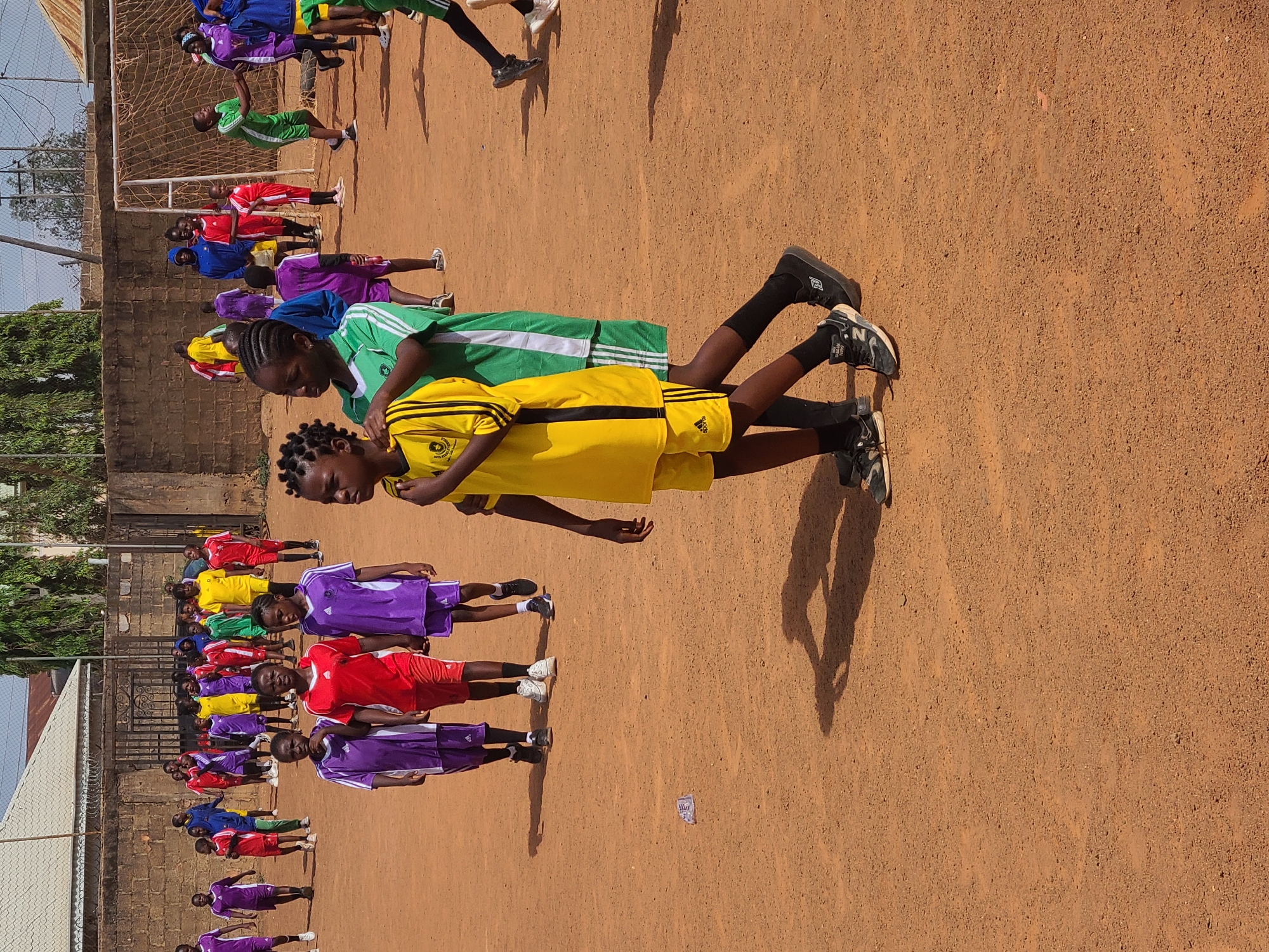 On a clear day in Barakin Lerawi, Nigeria, a group of children are assembled on a brown, dirt field adjacent to a building with a mesh-like fence. The children are lined up and paired, apparently stretching one another's backs. They wear assorted sports uniforms in colors of green, yellow, red, blue, and purple. The background includes a net structure and trees. The sun casts a distinct shadow of a pair of people onto the field. A piece of trash lies on the ground.