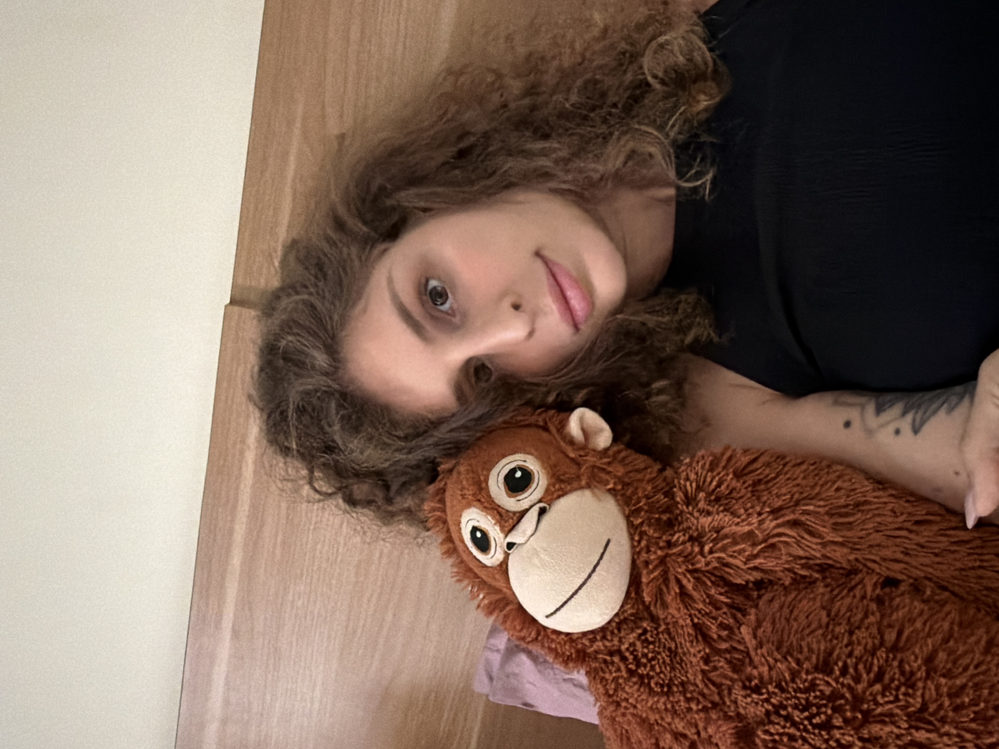 A woman with curly brown hair is lying in bed with a plush orangutan toy. She is wearing a black sleeveless top. The bed has a wooden headboard and a purple pillow. The room is dimly lit. The image was taken in North Kuta, Indonesia.