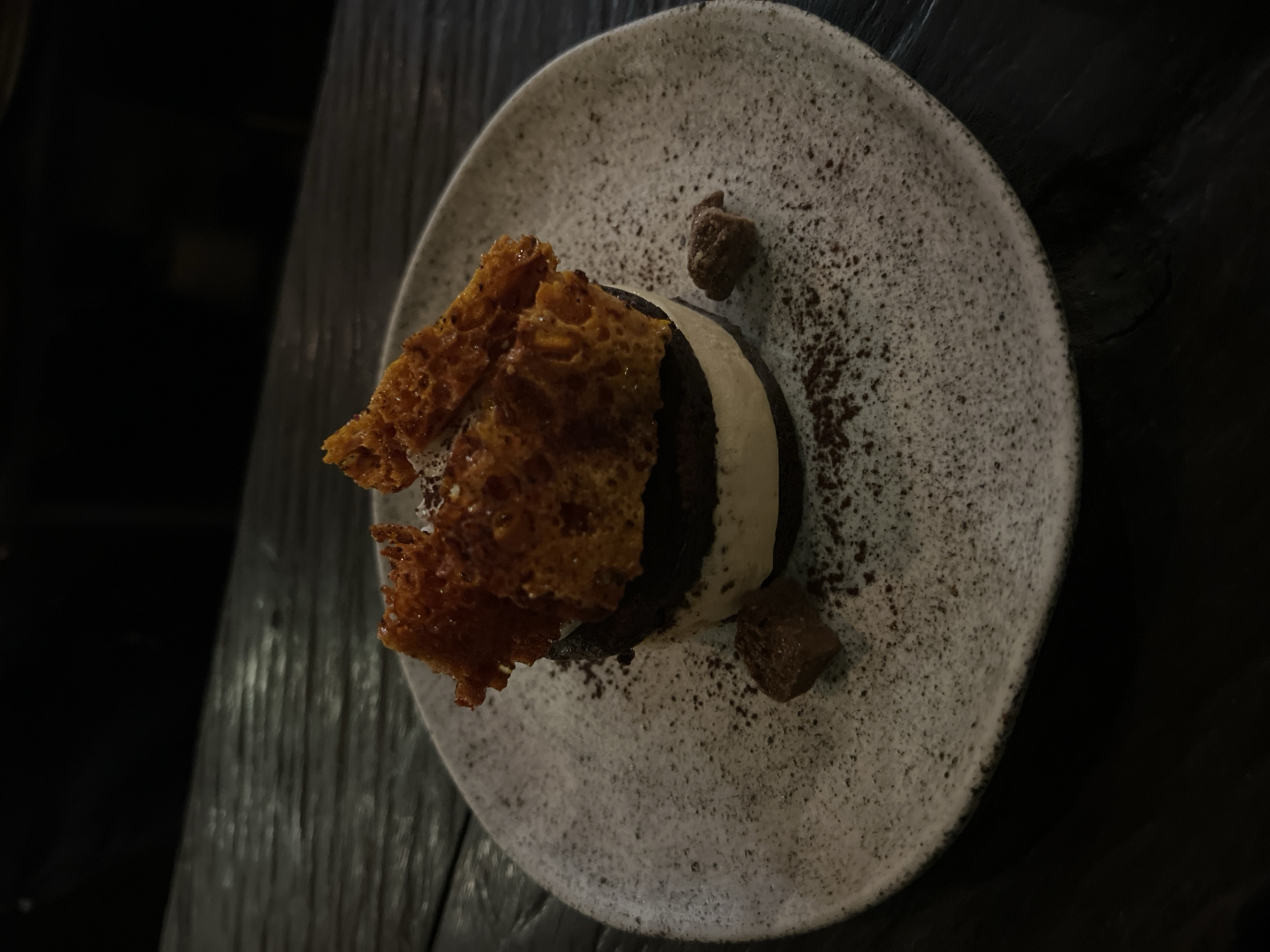 The image depicts a dessert on a plate. The dessert consists of what appears to be a layered element, featuring dark chocolate and light cream, topped with brittle pieces. Two small chocolate pieces are scattered nearby and there is cocoa powder on the plate. The plate is textured, and its edges are visible. The image is shot from an elevated angle, with a dark background and wooden surface in the lower-left corner. The lighting is subdued. It appears to be a restaurant setting in Mengwi, Indonesia. There are no people visible.