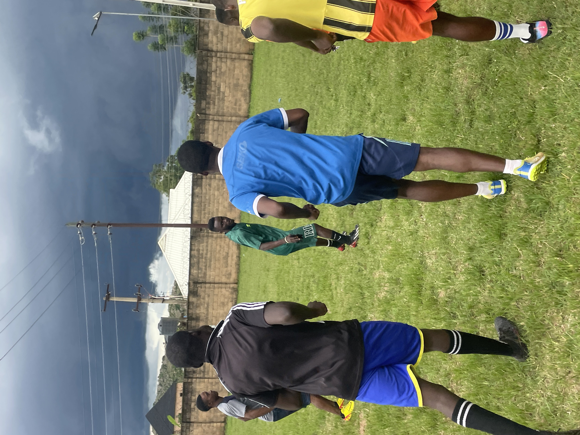 A group of young Black men in athletic wear are on a grassy field, possibly for a soccer practice, under a dramatic, dark, cloudy sky in Jos, Nigeria. The man in the foreground on the left wears a black and white striped shirt and blue shorts with yellow trim, black socks with white stripes, and black soccer cleats. To his right, a man in a blue shirt with white trim and dark shorts runs ahead. Further back, a man in a green shirt and dark shorts is visible. On the far right, a man in a yellow and black striped shirt and orange shorts is also running. The field is surrounded by a light brown brick wall topped with barbed wire. Beyond the wall, houses with varied rooflines and trees are visible. Power lines and utility poles crisscross the sky. The overall impression is of a sports activity taking place in an urban or peri-urban environment in Jos, Nigeria, with a stormy weather system approaching.