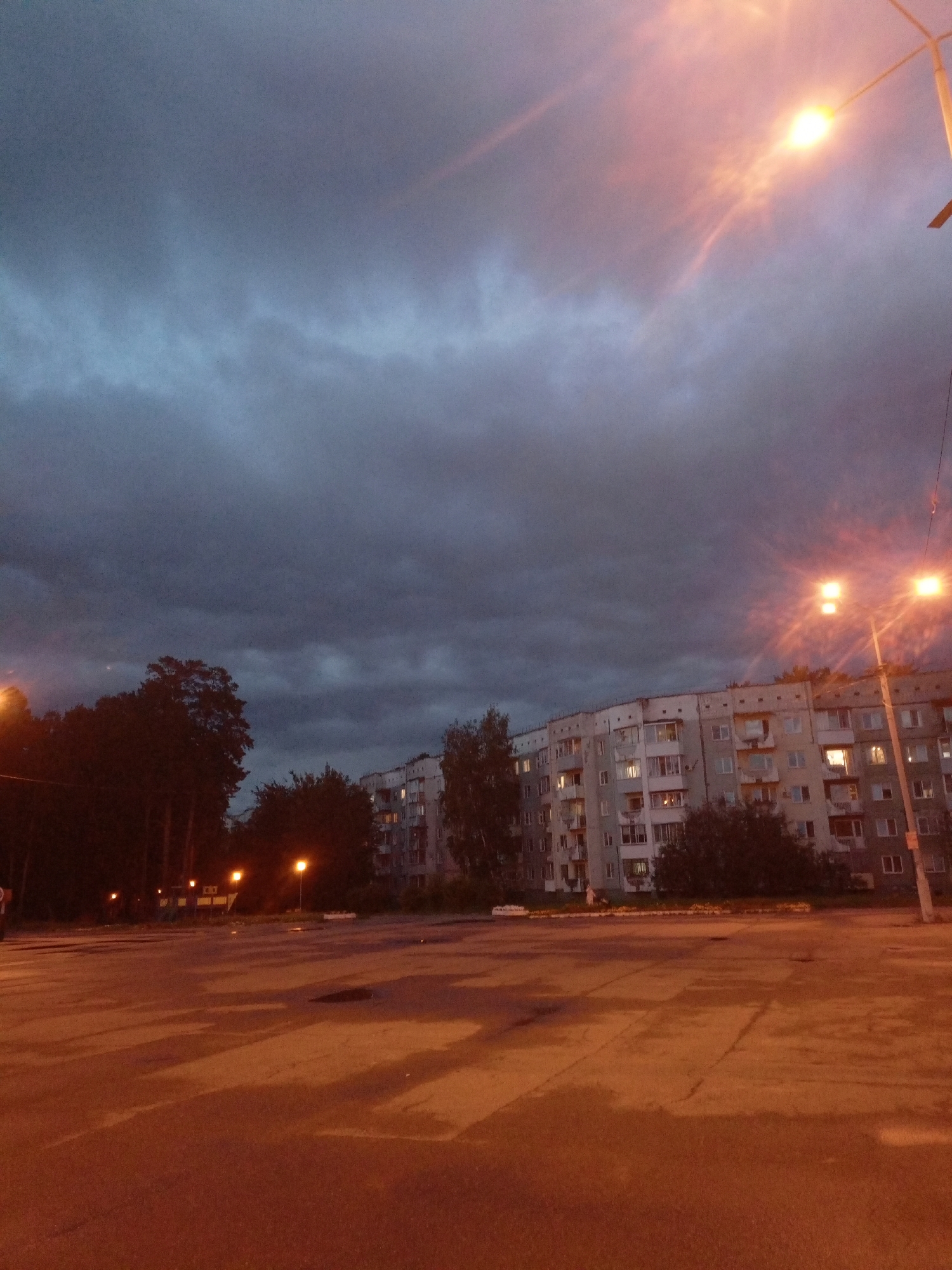 The image depicts an evening scene in Zheleznogorsk, Russia, likely a residential area. In the foreground, a paved surface, possibly a parking lot or square, exhibits reflections from streetlights, indicating it may be wet. The middle ground features a multi-story apartment building with illuminated windows, suggesting occupancy. Adjacent to the building are several trees, silhouetted against the darkening sky. A streetlamp with two illuminated bulbs is prominently visible in the upper right quadrant of the image, casting a warm glow. Another streetlamp is visible on the left side of the image, further from the viewer. The sky is overcast with heavy, dark clouds, tinged with a pinkish-orange hue, possibly from the setting sun or artificial lighting. The overall atmosphere suggests dusk or the onset of nightfall. No people are visible in the scene.
