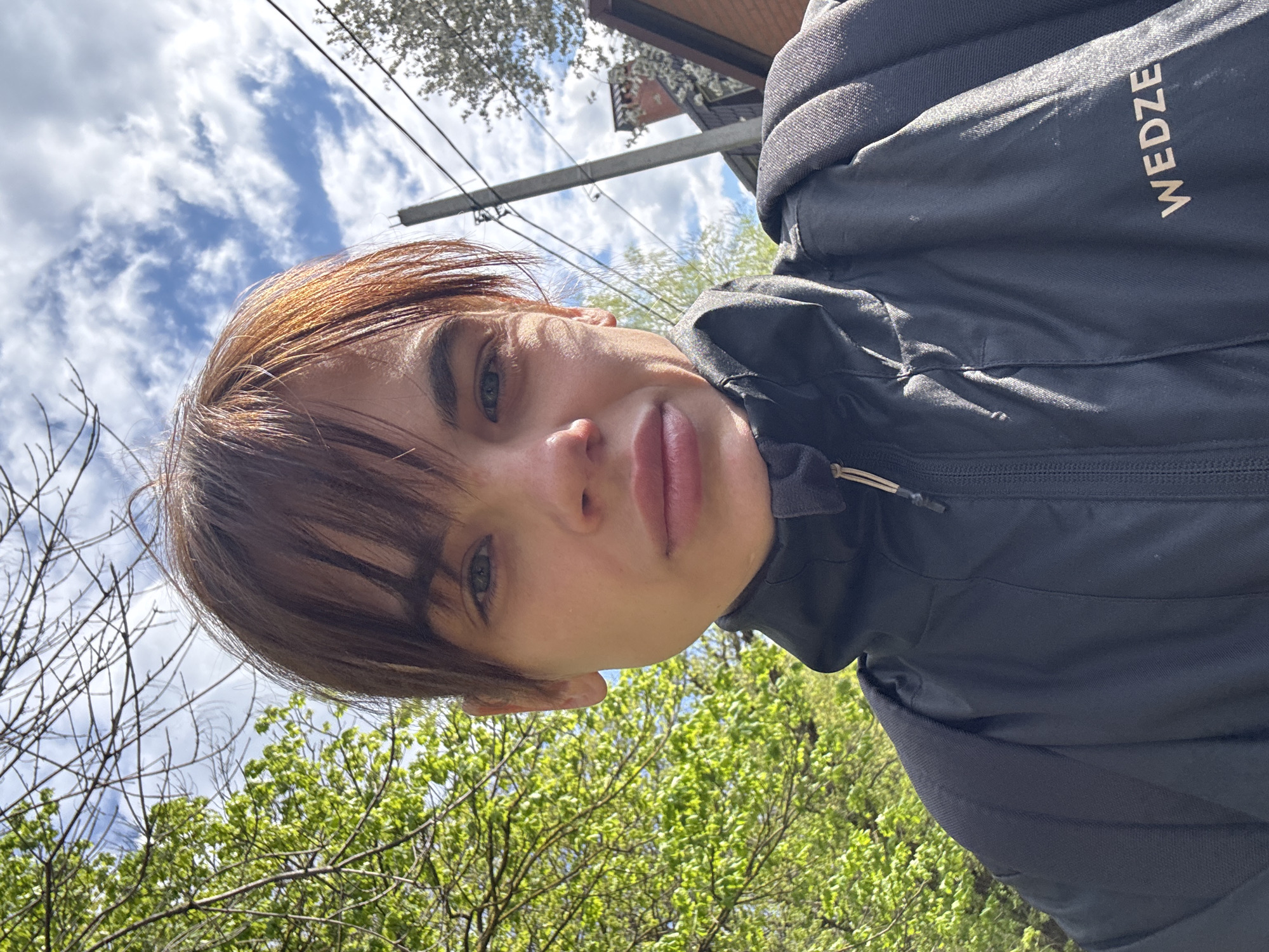 A close-up selfie shows a person with short, reddish-brown hair and bangs, with their face angled towards the camera. They have light-colored eyes and full lips, displaying a neutral expression. The individual is wearing a dark grey or black zippered jacket, with the brand name "WEDZE" visible in white text on the upper right side of the garment.
The background, predominantly on the left side of the frame, features a bright blue sky with scattered white clouds, suggesting a clear day. On the right, lush green trees with fresh leaves are visible, indicating spring or early summer. Some bare branches are also present. A utility pole with wires stretches horizontally across the upper middle portion of the background, and a building with a reddish roof or facade can be partially seen behind it. The scene is set outdoors in bright daylight. The image appears to be taken from a low angle, looking upwards.
