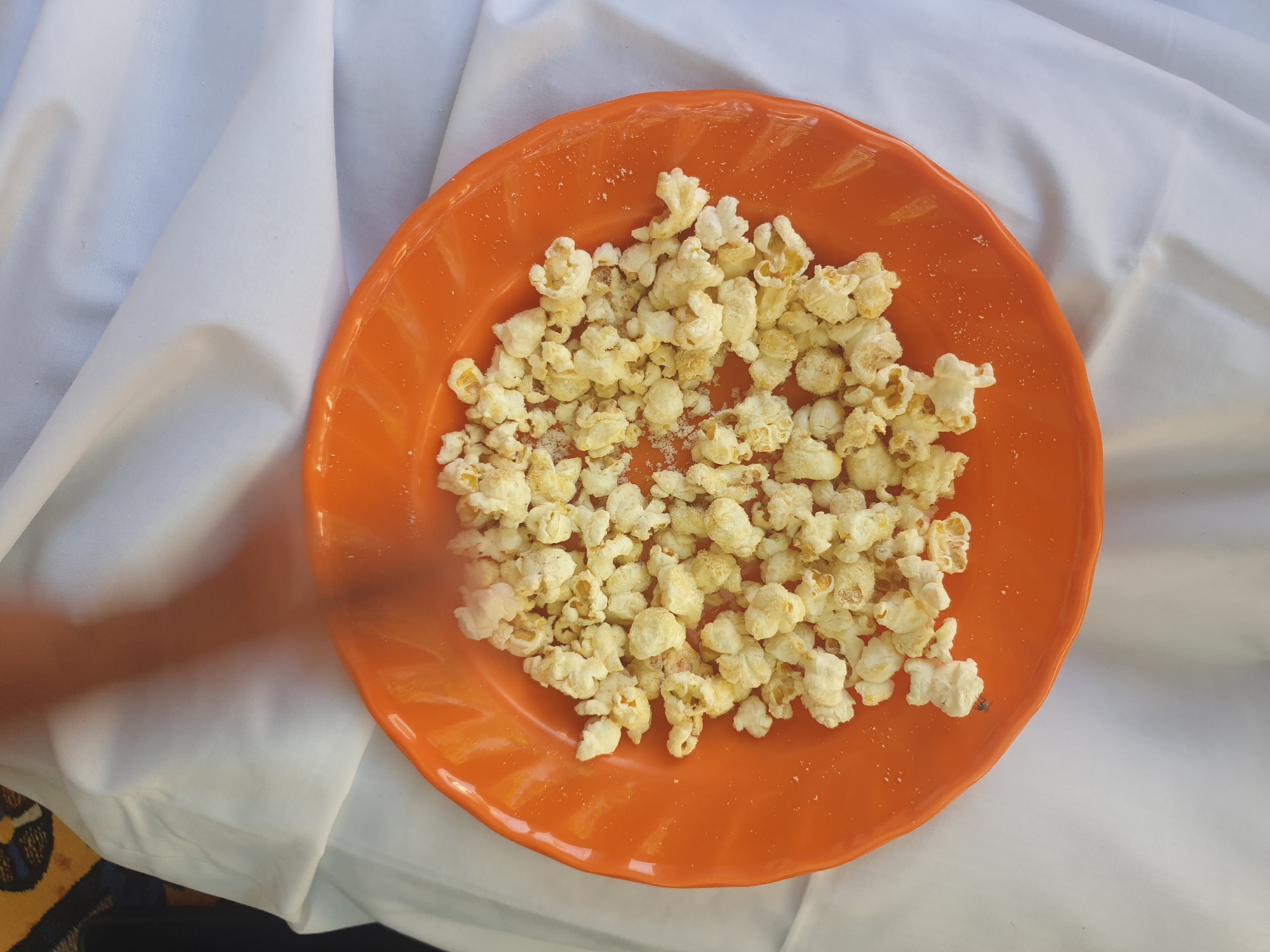 The image displays an overhead view of a vibrant orange, round plate filled with plain, white popcorn, lightly seasoned with visible salt granules scattered across both the kernels and the plate's surface. The plate rests on a white, rumpled fabric surface, likely a tablecloth or bedsheet, which covers most of the background. In the upper-left corner, a glimpse of a darker, patterned fabric, possibly a rug or blanket with blue and yellow designs, is visible beneath the white cloth. A blurred human arm, with light brown skin, is captured in motion from the top of the frame, reaching towards the popcorn, indicating an interaction with the snack. The lighting is bright and even. The scene is situated in Jalingo, Nigeria.