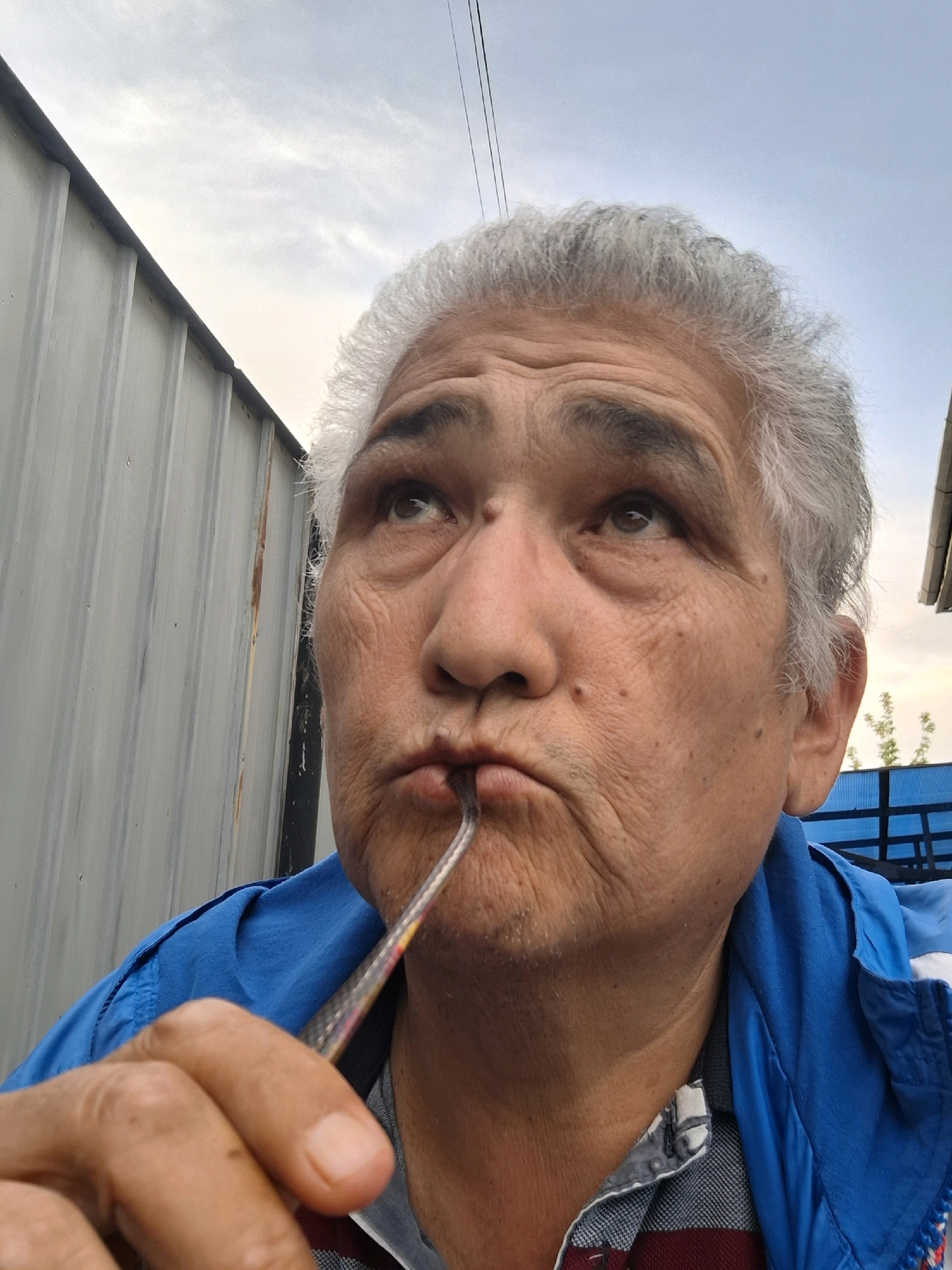 The image shows a close-up, eye-level, outdoor selfie of an elderly person with gray hair. The person is wearing a blue jacket over a checkered shirt and has a utensil in their mouth. They are looking upwards and to the right. The background consists of a corrugated metal wall on the left, a blue tarp structure on the right, and a pale sky with some clouds and power lines. The scene appears to be in an industrial or utilitarian area, possibly a yard or alleyway. There are no specific landmarks visible to pinpoint the location within Almaty, Kazakhstan.