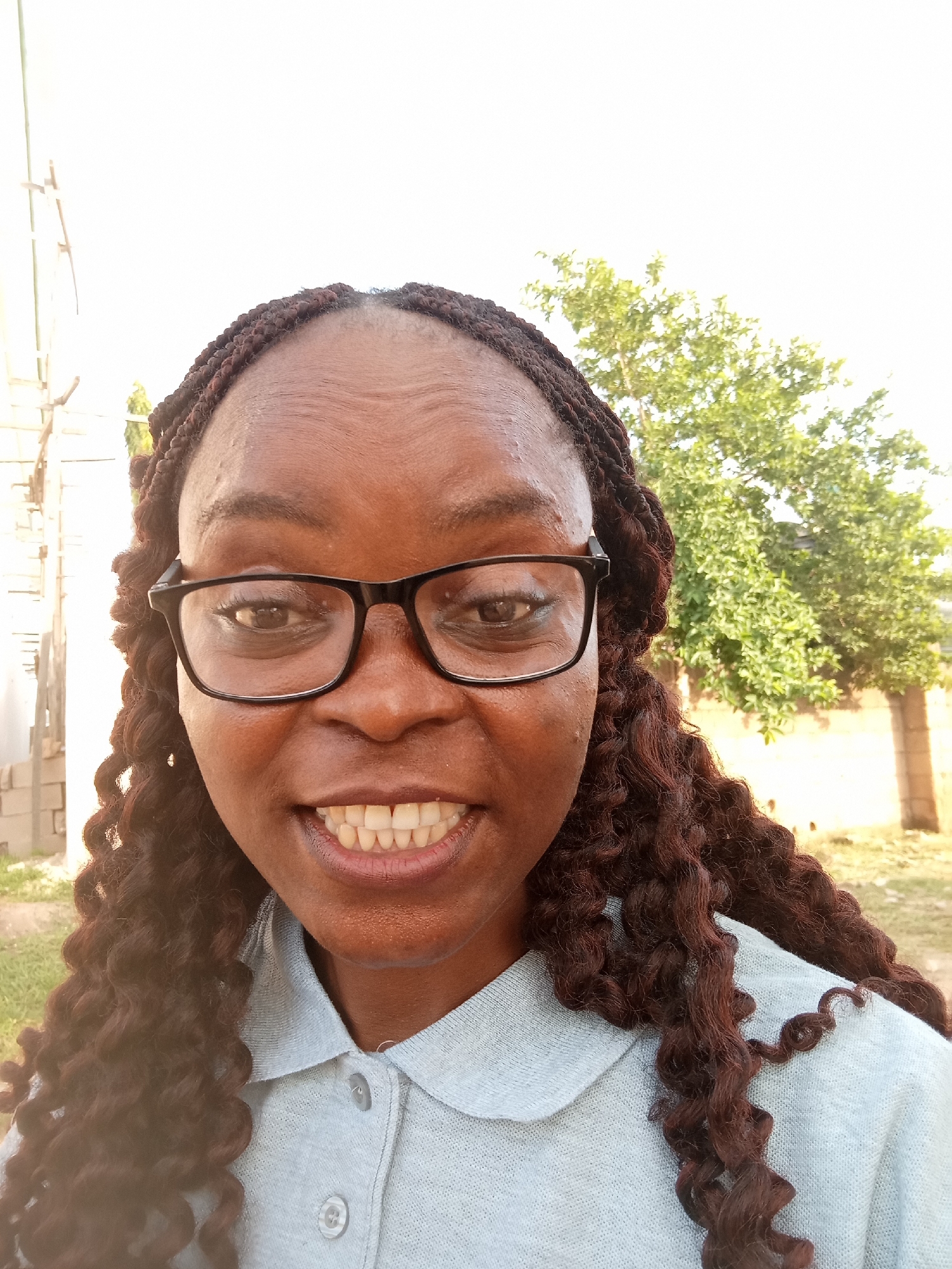 A medium-shot photograph shows a dark-skinned woman wearing black-rimmed glasses and a light blue collared shirt. Her hair is styled in dark brown braids that fall around her shoulders, with some curly extensions. She is smiling, displaying a row of white teeth. The background is slightly blurred, with a pale sky and green foliage from a tree. To the left, a light-colored building with vertical elements is visible, and to the right, a tan wall and some dry grass. The lighting suggests it is daytime, possibly outdoors. This scene appears to be in Jalingo, Nigeria, an area characterized by its warm climate and open landscapes.