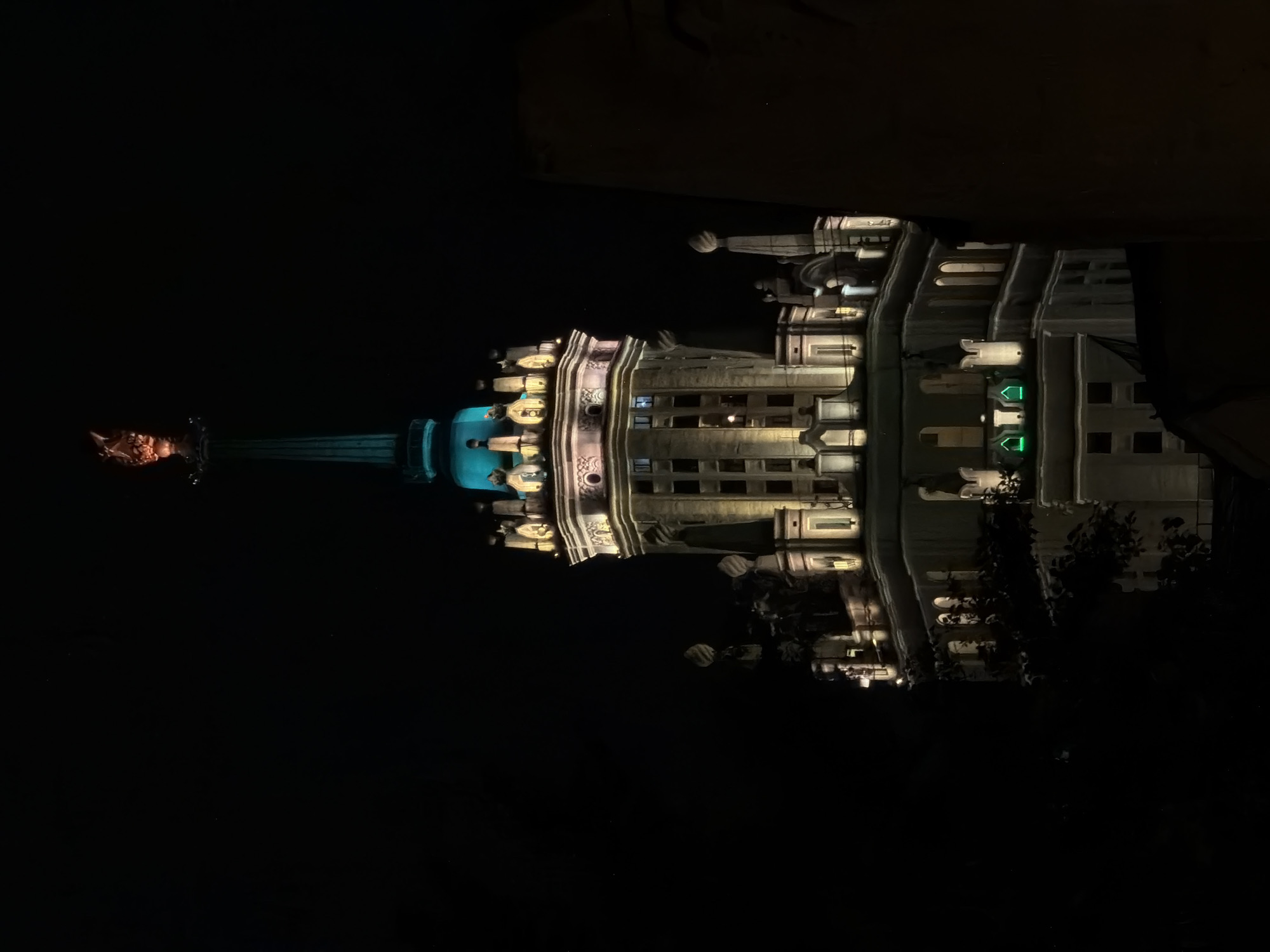 At night, a building with a tall spire is brightly illuminated. The building has a classical architectural style, with decorative elements and rows of windows. The spire is topped with an illuminated star-like structure. The image appears to show a low angle of the building. The scene is in Moscow, Russia.