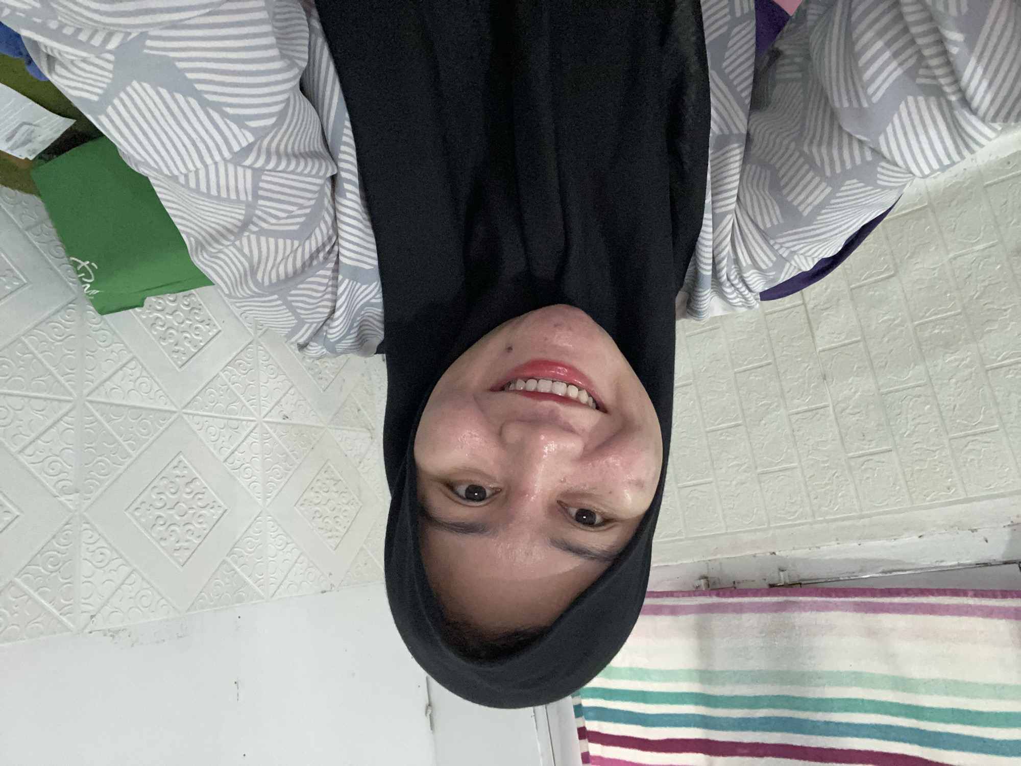 In Banjarbaru, Indonesia, a close-up, frontal selfie shows a young woman wearing a black hijab and a grey and white patterned shirt. She has a bright smile, revealing her teeth, and appears to have red lipstick on. Her skin shows some texture and shine. The background consists of a textured white wall and a striped fabric hanging on the left. A green bag is visible on the right side of the image.