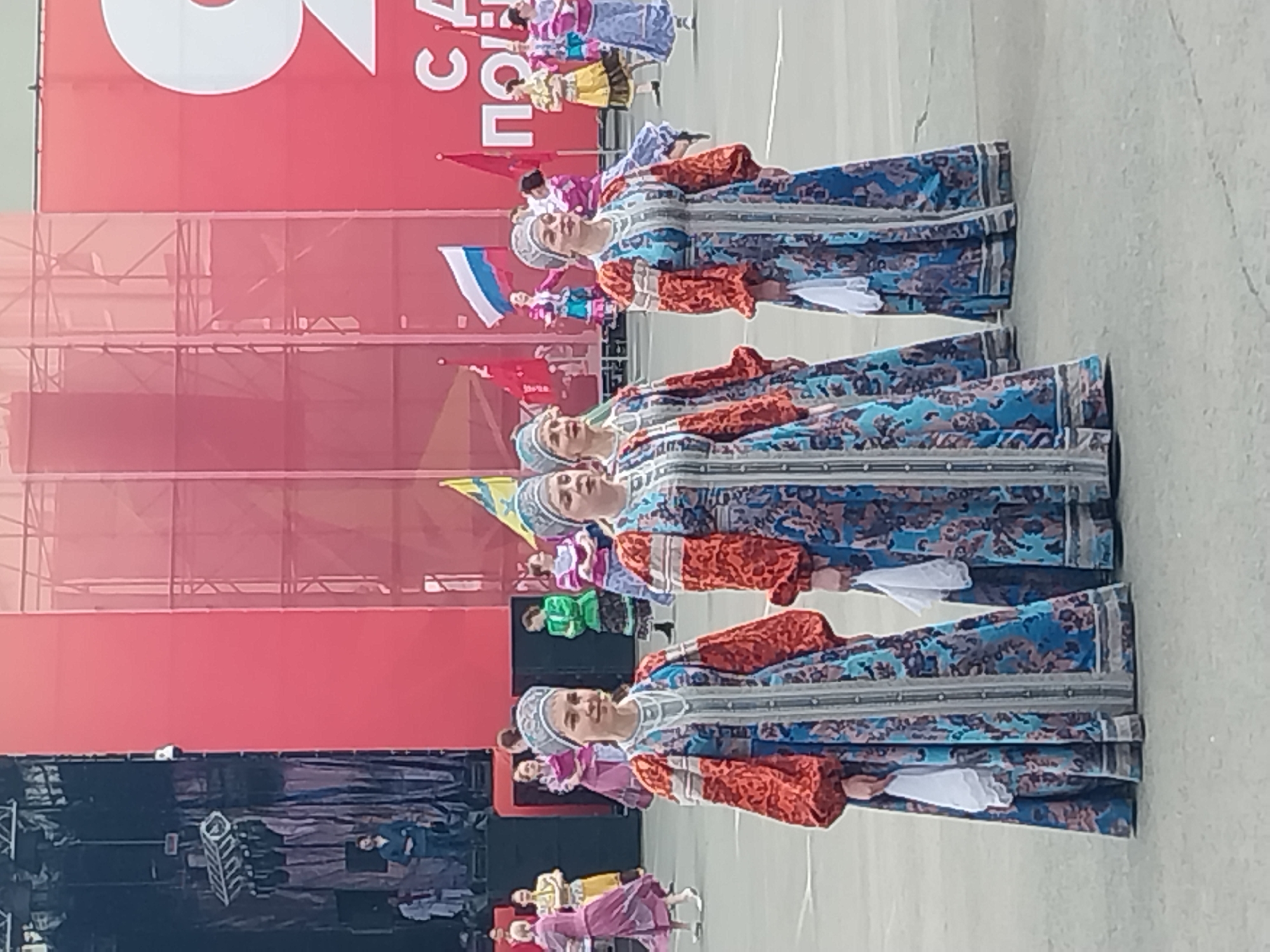 Four women in traditional Russian attire are standing in a line, facing forward, likely for a performance or celebration. They are wearing long, patterned blue and red dresses with white head coverings and holding white cloths. Behind them is a red wall with white lettering and flags. There are other people in the background in similar, yet less elaborate, traditional clothing. The ground is a light-colored, paved surface. It is daytime. The location is most likely in the city of Orenburg, Russia.