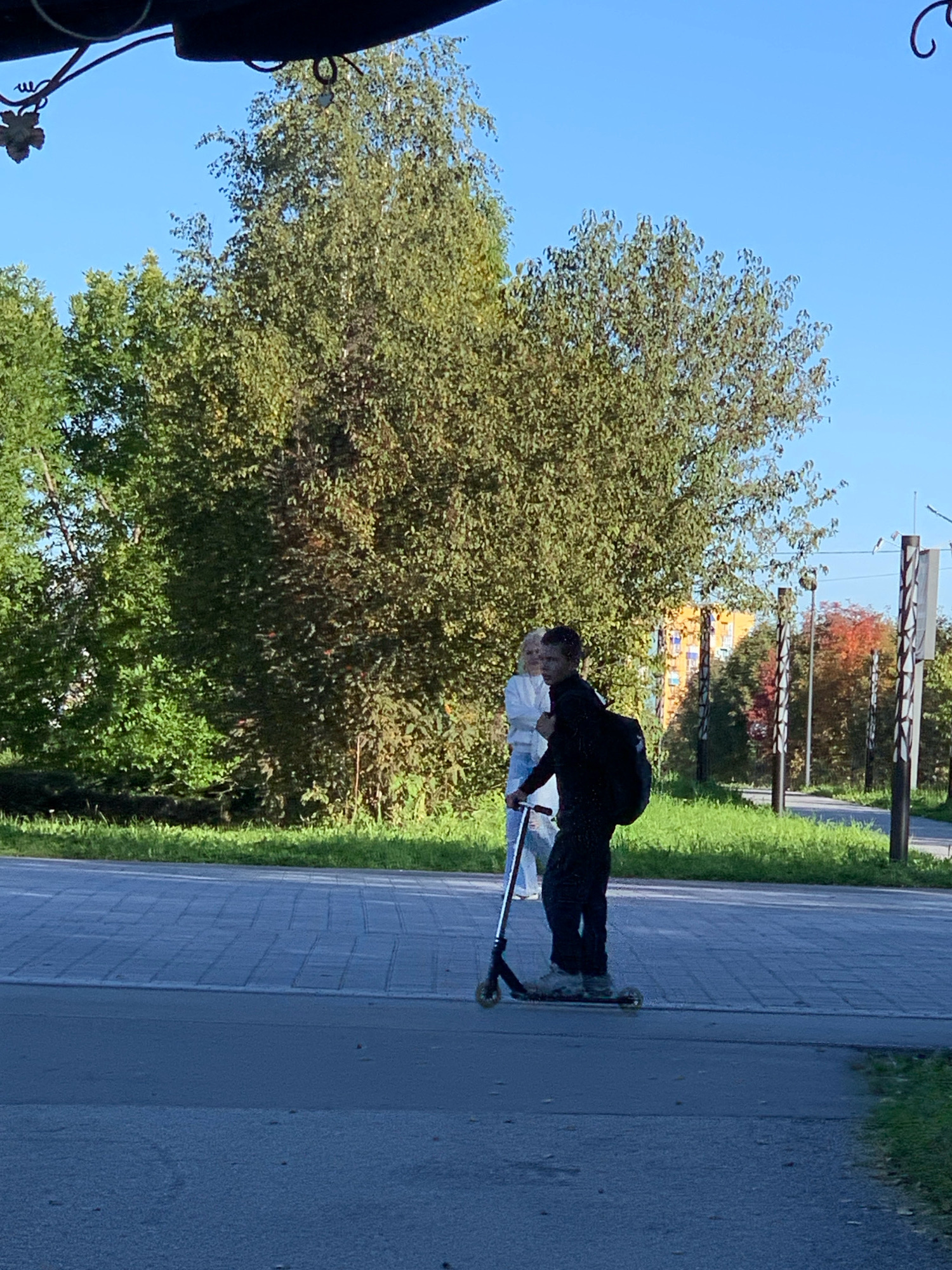 In Prokopyevsk, Russia, on a paved walkway next to a grassy area, a young male wearing dark clothing and a backpack is riding a scooter. To his left, a young female in light-colored clothing stands on a scooter. Behind them, mature trees with green foliage dominate the background, with an orange building visible in the distance. The sky is clear and blue.