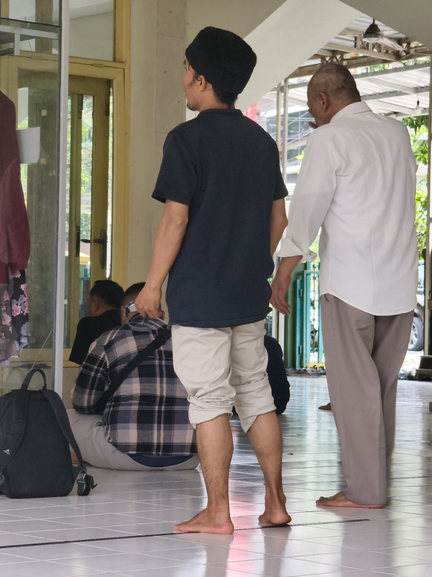 The image depicts an outdoor setting in Samarinda, Indonesia, likely a communal area near a place of worship. In the foreground, two men are the primary subjects. The man on the left is wearing a dark beanie, a dark short-sleeved shirt, and light-colored shorts. He is barefoot and facing away from the camera, looking towards the right. To his right, an older man in a white long-sleeved shirt and grey trousers is also facing away from the camera, with his right hand near his mouth. He appears to be speaking or gesturing.
Behind these two men, a third man is partially visible, seated and wearing a plaid shirt. He is also barefoot. Further in the background, through an open doorway or window, a reflection shows another man wearing a dark shirt.
The environment features a tiled floor with a grid pattern. To the left, a large black backpack rests on the floor near a doorframe with yellow accents and glass panels. The background suggests an open-air structure with a roof supported by metal beams, and greenery is visible beyond the immediate area, indicating an outdoor or semi-outdoor space. The lighting suggests it is daytime.