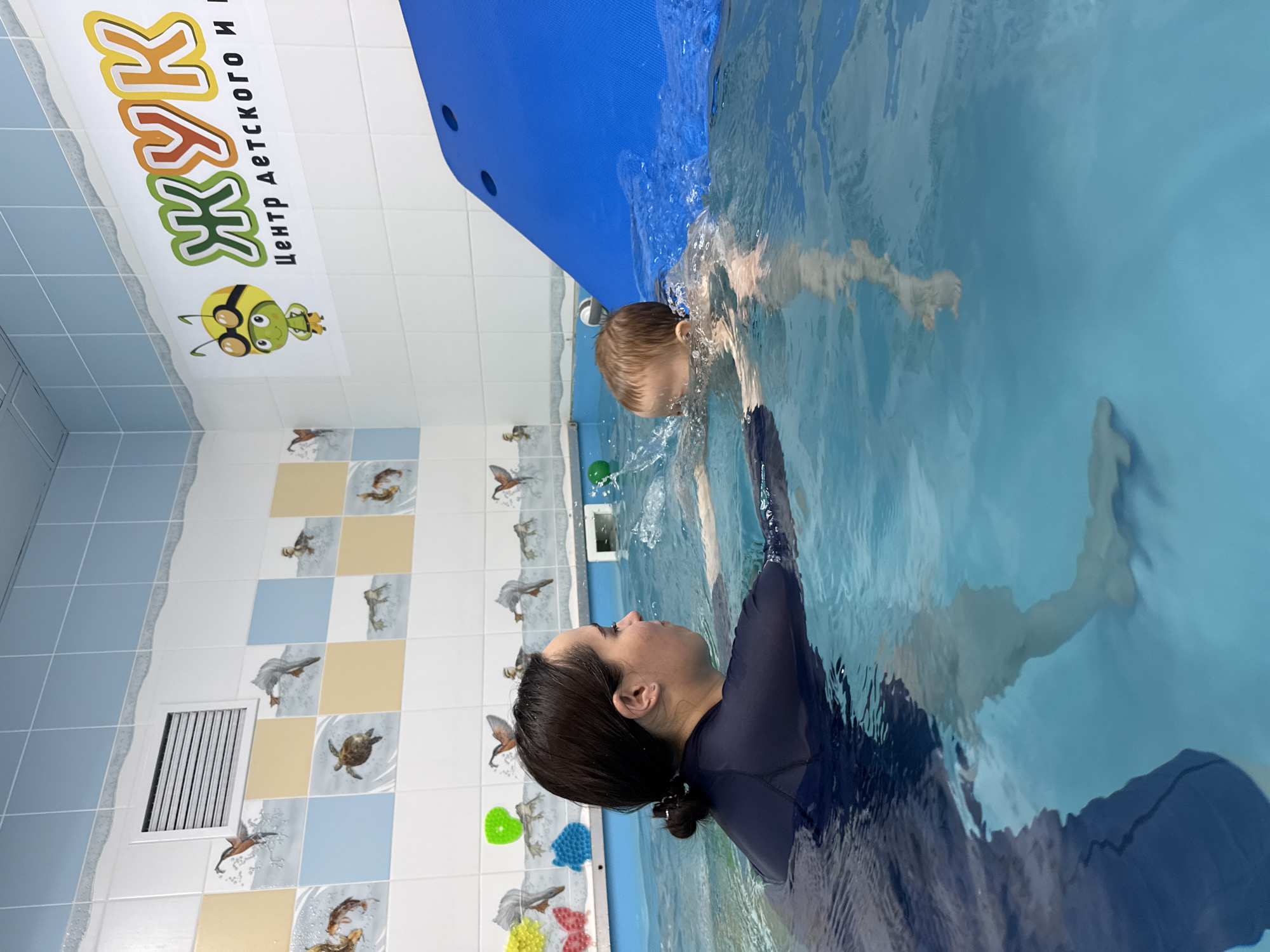 A woman in a navy blue rashguard is in a swimming pool with a child. The child is submerged in the water, and the woman appears to be supporting or assisting the child.
The pool is located in what appears to be an indoor facility, with tiled walls decorated with images of aquatic life, birds, and sea turtles. There is a sign in Russian on the wall, as well as blue and green floating toys near the edge of the pool.
The location in the city of Rusanovo, Russia is not directly identifiable from this image, although the Cyrillic signage suggests the image was taken in Russia.