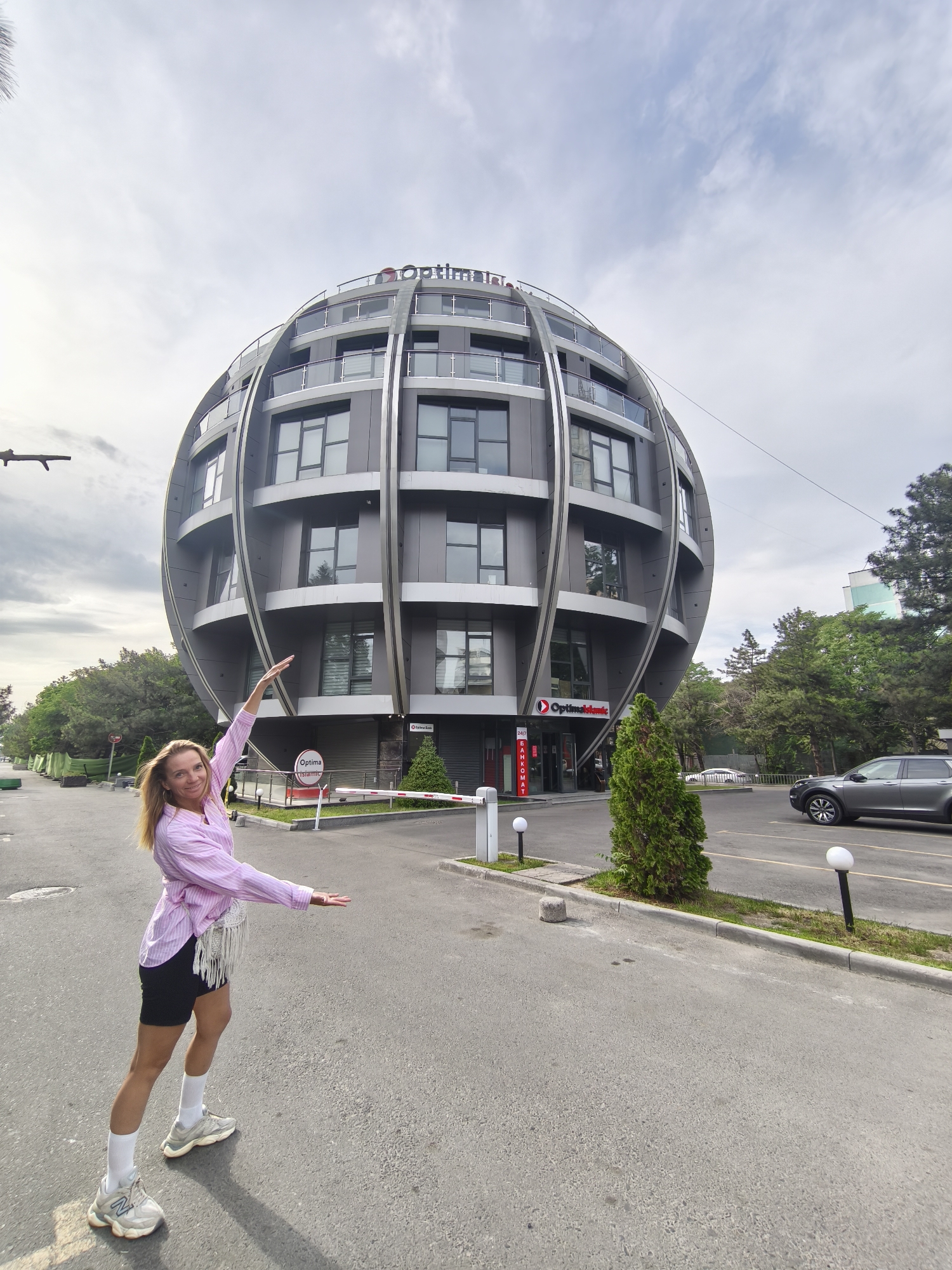 The image shows a blonde woman standing in a street in front of a distinctive spherical building. The building, identified by the "Optimal Bank" signage, is located in Bishkek, Kyrgyzstan. The building has multiple floors with windows and balconies, encased by metal framework.
The woman is wearing a pink and white striped shirt, black shorts, white socks, and running shoes. She has her arms outstretched, gesturing towards the building. Behind her, a barrier arm is in place near the entrance of the parking lot of the building. A grey SUV is parked nearby. Trees surround the scene, and the sky is overcast. The street appears to be paved and clean.