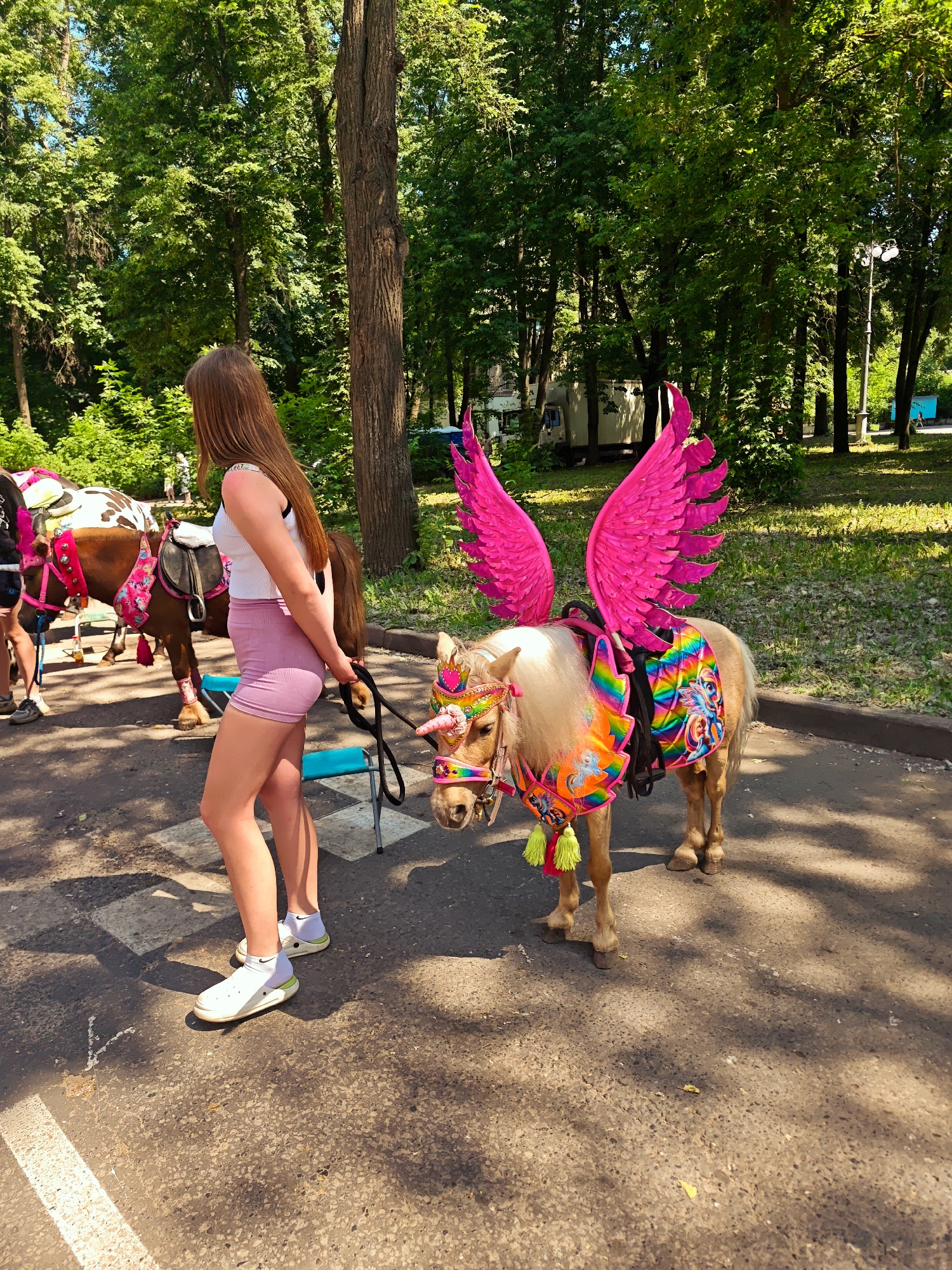In a park setting in Kimry, Russia, a young woman with long brown hair, wearing a white tank top and pink shorts, stands to the left of a miniature horse. The horse is adorned with bright pink wings, a rainbow-colored saddle pad, and a unicorn horn on its forehead. The woman holds a lead rope attached to the horse's bridle. Another pony, decorated with pink and white accents, is visible behind them. The ground is paved with asphalt, and lush green trees line the background under a bright sky. The scene appears to be a festive event or fair.