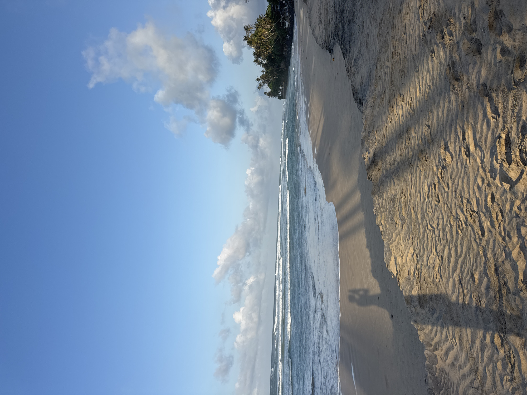 This is a photograph of a beach in Kathaluwa, Sri Lanka. The image shows a sandy beach with gentle waves rolling in from the ocean. Palm trees and lush vegetation line the coastline in the distance. The sky is a clear blue with a few scattered clouds. The foreground of the image features the texture of the sand, with ripples and footprints visible. A shadow of a person holding a camera is cast on the sand, suggesting the photograph was taken by someone at the beach. The overall scene conveys a sense of tranquility and natural beauty.