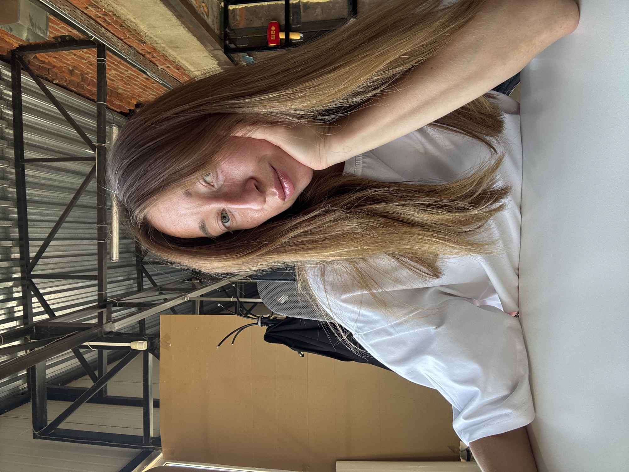A woman with long brown hair is sitting at a white table indoors, resting her head in her hand. She is wearing a white t-shirt. Above her, the ceiling is constructed of metal supports and corrugated metal sheets, typical of industrial or commercial buildings. Behind her, there is a brown wall, a coat hanging on a coat rack, and a mesh office chair. A possible location could be a workplace or an industrial building in the city of Chaikovsky, Russia.