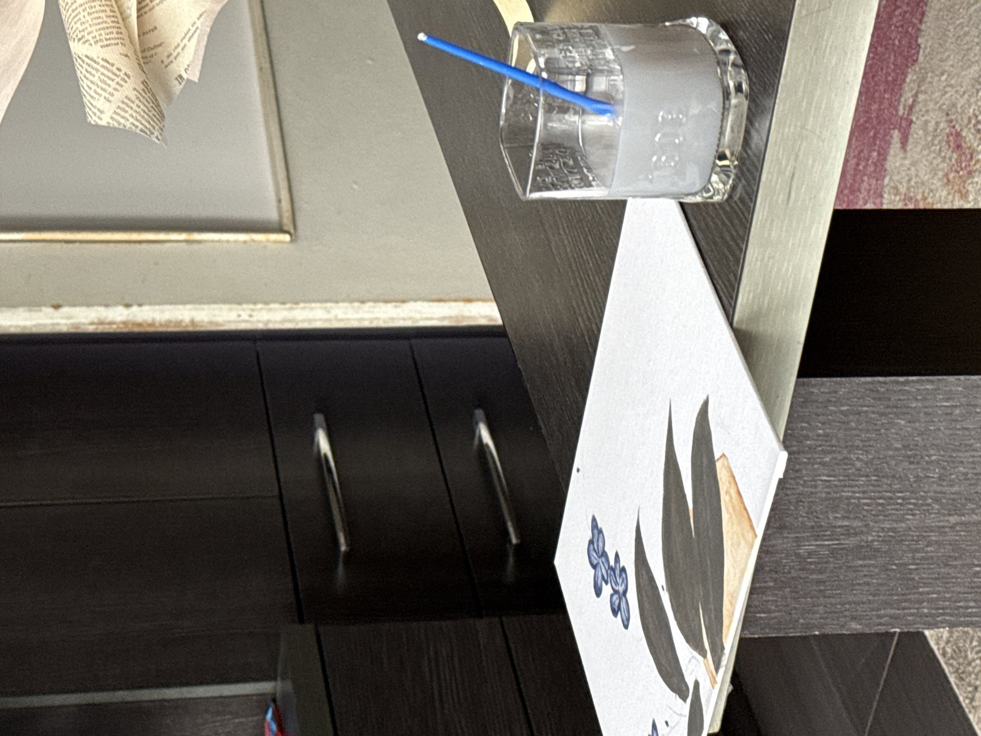 The image shows a close-up of furniture and art supplies. A clear glass with a blue straw stands on a dark gray surface, next to a piece of white cardstock with a floral design. A brown cabinet with silver handles is visible in the background. The exact location in Sofia, Bulgaria, cannot be determined from the image.