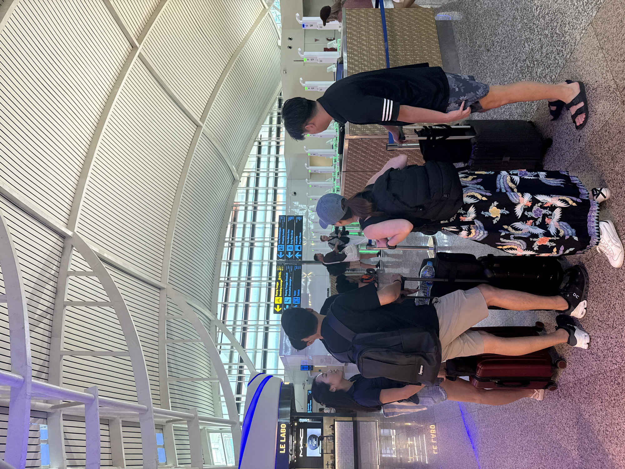 In the departure hall of Ngurah Rai International Airport in Kuta, Bali, Indonesia, a group of five individuals stands near a check-in counter area. Two women and three men, all appearing to be tourists or travelers, are gathered with their luggage. One woman, wearing a dark blue polo shirt and denim shorts, faces left, engaged in conversation. Beside her, a man in a black t-shirt and light brown shorts faces away from the camera, holding a rolling suitcase. Another man, also in a black t-shirt and patterned shorts, stands with his back to the viewer, his hand resting on a black suitcase. A second woman, dressed in a black and white patterned skirt and a blue baseball cap, faces left. The fifth individual, a man in a black t-shirt and patterned shorts, stands to the right, also facing away from the camera, with a black rolling suitcase.
The environment is an airport terminal with a high, modern ceiling structure featuring white, slatted panels and exposed beams. Fluorescent lighting illuminates the space, creating a bright atmosphere. In the background, digital signs display flight information in both Indonesian and English. A store labeled "LE LABO" is visible to the left, and a check-in counter with multiple stations, some displaying green lights, is situated further back. The floor is a speckled granite material. The overall scene depicts a typical airport scene of people preparing for departure.
