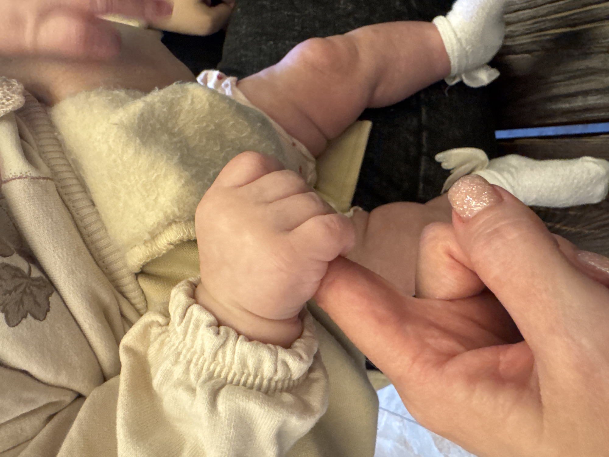 This close-up, high-angle indoor photograph captures an intimate moment between an adult and a baby. The primary subjects are the baby's tiny hand grasping the adult's index finger. The baby's left hand is centrally positioned, clad in a cream-colored, long-sleeved garment with a ruched cuff, holding onto the adult's finger.
The baby is dressed in several layers of light-colored clothing, including what appears to be a fuzzy, light-yellow or cream-colored inner layer and a patterned outer garment. Parts of the baby's torso, arm, and legs are visible; the legs show natural baby fat folds. The baby's feet are covered with white socks or booties that appear to be tied around the ankles.
The adult's right hand is visible on the right side of the frame, with the index finger being held by the baby. The adult's thumb and a portion of the palm are also visible, showing a manicured fingernail with glittery, light pink or nude polish.
The activity depicted is the common, tender interaction where a baby clutches an adult's finger, signifying connection, affection, and bonding. The scene evokes feelings of warmth, love, and protection.
The setting appears to be a cozy indoor environment, possibly a home in Chaikovsky, Russia. In the background, a dark, possibly wooden, surface is visible, along with dark fabric which could be clothing or a blanket. The lighting is soft and ambient, characteristic of an indoor shot. There are no visible location cues within the image itself to confirm a specific Russian location, nor any visible text. The time of day is not discernible.
