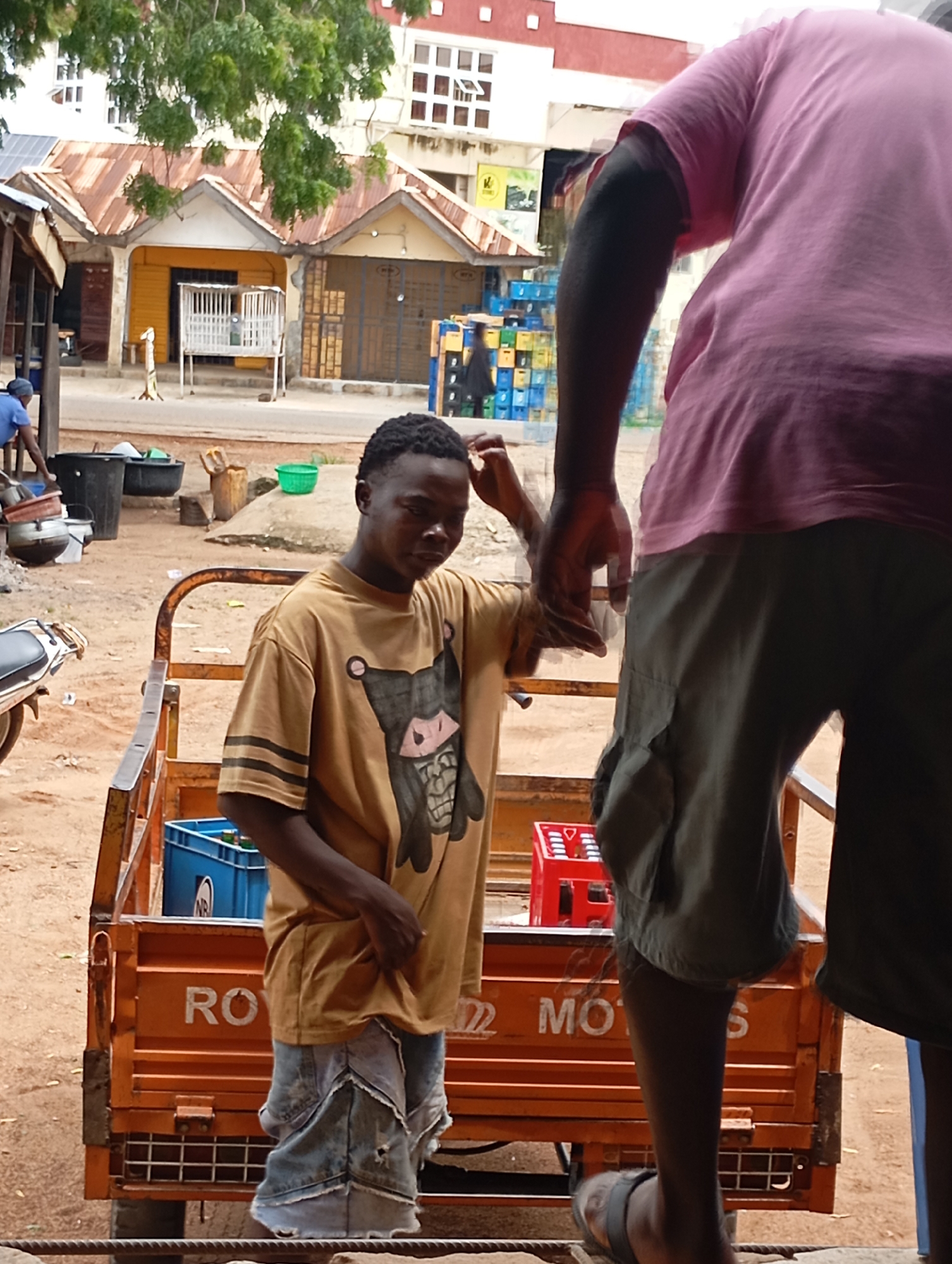 A young man wearing a light brown t-shirt with a black and white cartoon character on it and ripped denim shorts stands next to an orange utility cart. The cart has the word "ROY" and "MOS" visible on its side. The man is holding his left hand up to his head, as if scratching it. In the background, a person in a pink t-shirt is partially visible, bending over. Behind them, a building with a corrugated metal roof and white windows is seen. The ground is dirt, and there are various objects scattered around, including plastic crates, buckets, and a green bucket. The scene appears to be an outdoor market or street scene in Shendam, Nigeria.