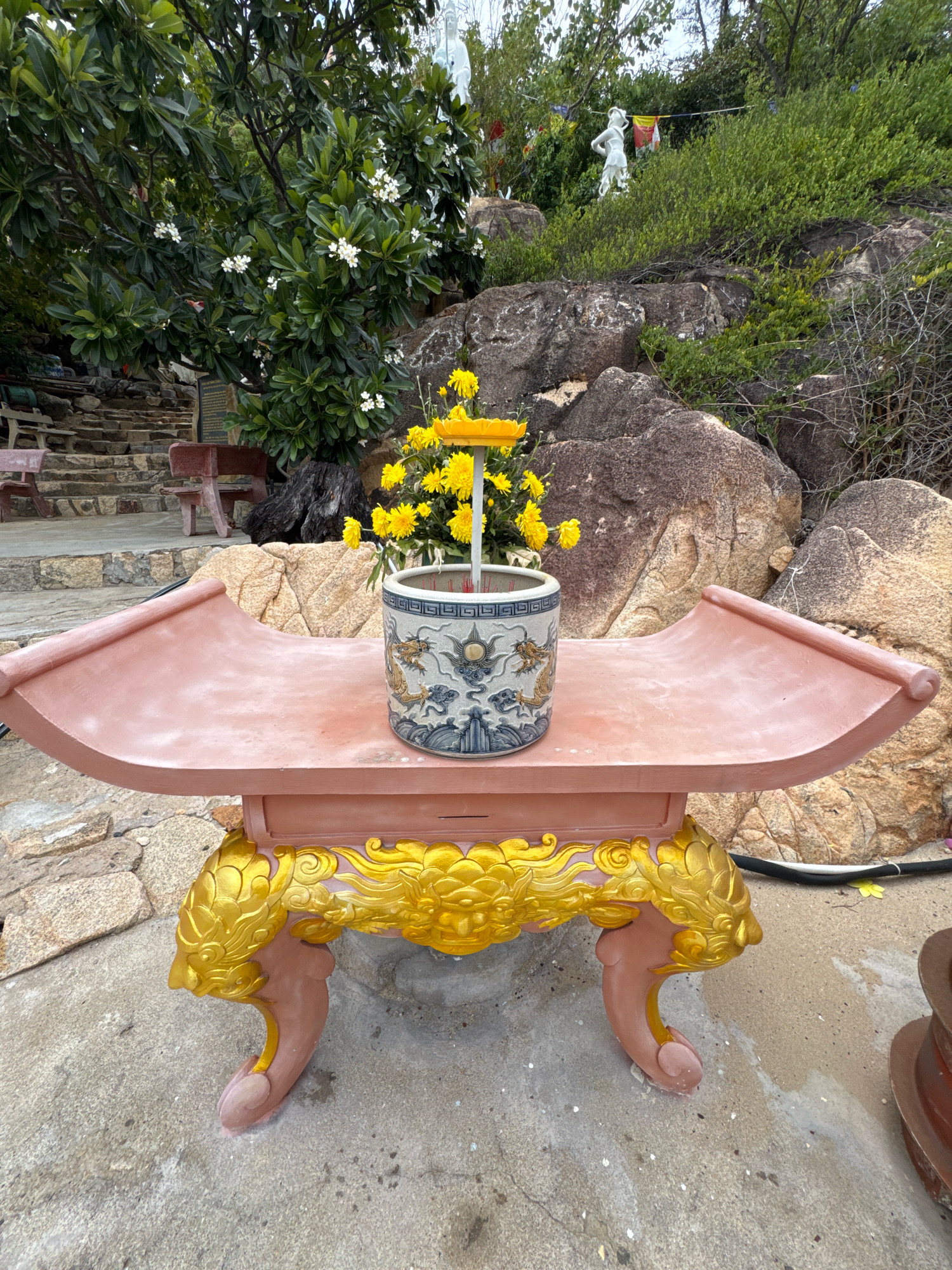 A close-up, eye-level, medium shot captures a decorative table with a vase of yellow flowers in the foreground, set against a backdrop of rocks and greenery. The table, with its curved, pinkish top and ornate golden dragon-like carvings on its legs, appears to be made of a faux stone or plaster material. It sits on a rough concrete surface.
Inside the vase, which is adorned with intricate blue and white dragon motifs, is a bouquet of bright yellow chrysanthemums, with a yellow stand holding them. Several red incense sticks are visible within the vase. The background reveals a rocky hillside covered with lush green vegetation. A stone staircase and benches are visible to the left, suggesting a public or spiritual site. Further up the hill, a white statue of a figure, possibly a deity or religious icon, stands amidst the foliage, with prayer flags or banners draped between structures. A portion of a large brown pot or container is visible on the far right. The lighting suggests it is daytime, with overcast or diffused light. The image is taken outdoors in what is likely a temple or spiritual garden setting in Nha Trang, Vietnam.