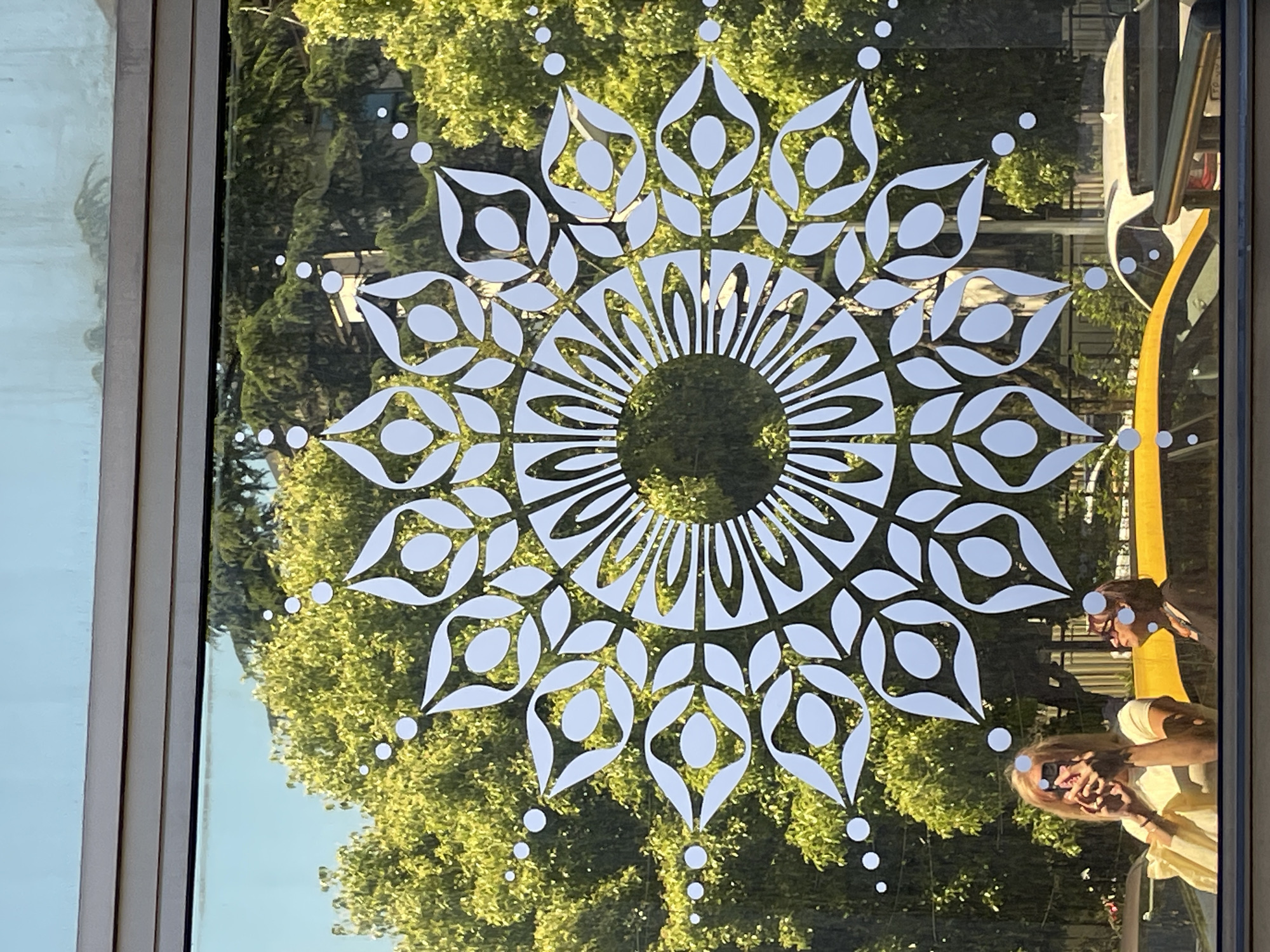 The media file presents a clear glass window adorned with a large, intricate white decal, viewed from the interior looking outwards. The decal is a symmetrical, mandala-like pattern featuring a central circular opening surrounded by concentric rings of petal or teardrop shapes, interspersed with small white dots.
The window's surface reflects the external environment, prominently displaying lush, dense green foliage from trees, indicating a vibrant outdoor setting under bright sunshine. In the reflections, parts of a building or structure are visible, along with a partial reflection of a vehicle, possibly white, featuring a distinct yellow section on the right side of the frame.
Two individuals are reflected in the lower right portion of the window. One person, likely the photographer, is seen wearing a light-colored top and sunglasses, holding a device (presumably a phone or camera) to take the picture. A second person's reflection is partially visible above and behind the first, appearing in a darker top.
The image conveys a daytime scene with sunny weather, as evidenced by the bright reflections and illuminated foliage. The lush greenery is consistent with the provided location context of Sochi, Russia. No visible text is present in the image.