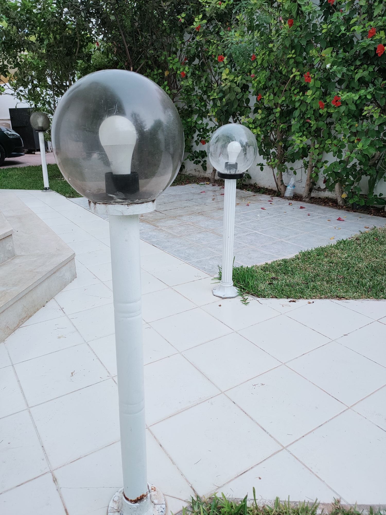 Two white-post lampposts with large, transparent spherical globes stand on a white tiled patio in front of a wall of green bushes with red flowers. The lampposts are of varying heights and are positioned at different distances from the viewer. The closest lamppost is in the foreground on the left, and the second lamppost is further back and to the right. The ground consists of large square white tiles, with a section of green grass and grey patterned tiles visible behind the lampposts. The background features lush green foliage and some red hibiscus flowers. The setting appears to be an outdoor space, possibly a garden or courtyard, in Mrezga, Tunisia.