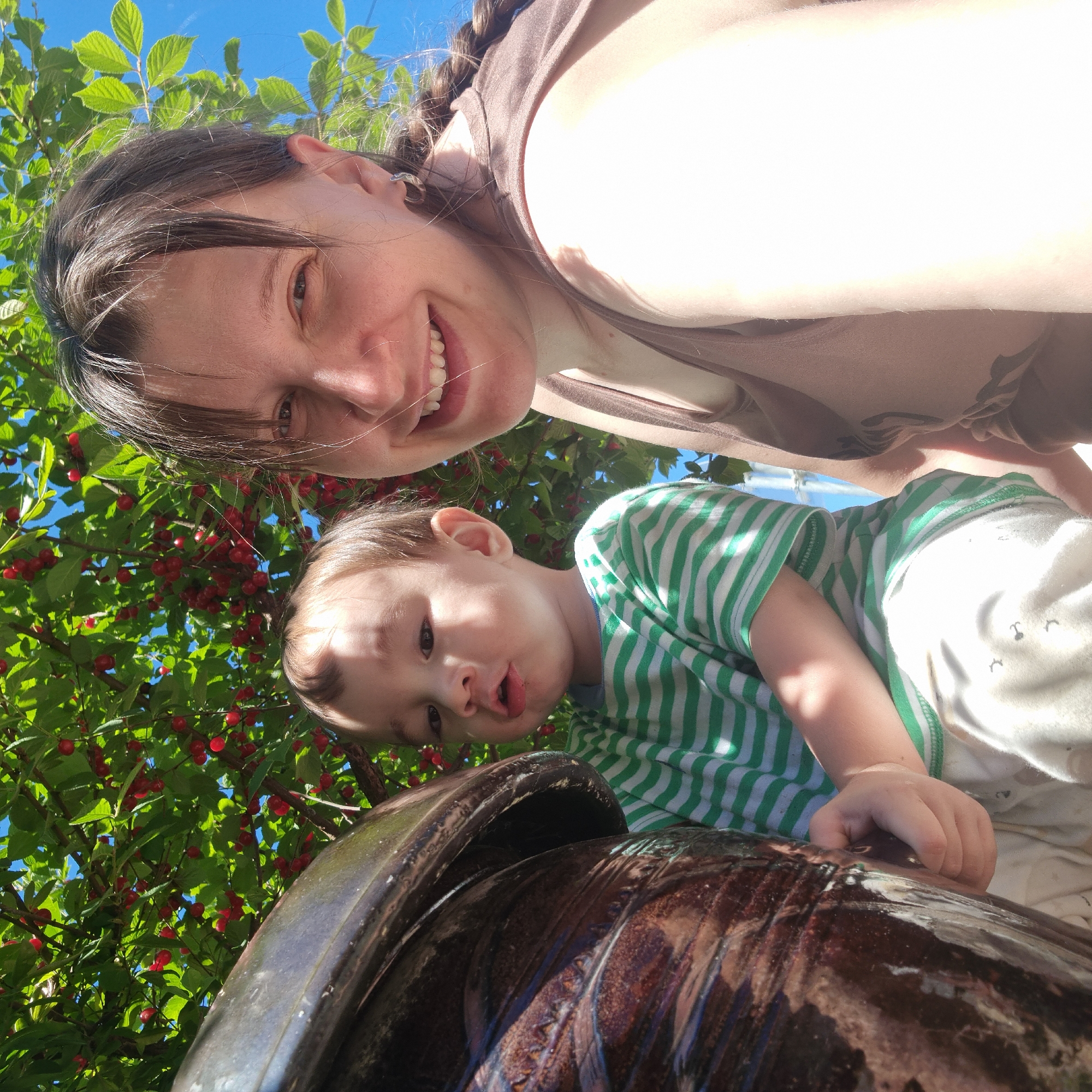 A woman and a young child are taking a selfie outdoors under a tree with red berries. The woman is smiling and looking at the camera. The child, in a green and white striped shirt, is looking at the camera with his mouth slightly open. A large, dark, textured pot is in the foreground. The background is a blue sky and green leaves with red berries. The scene is likely in Dunnae-myeon, South Korea.