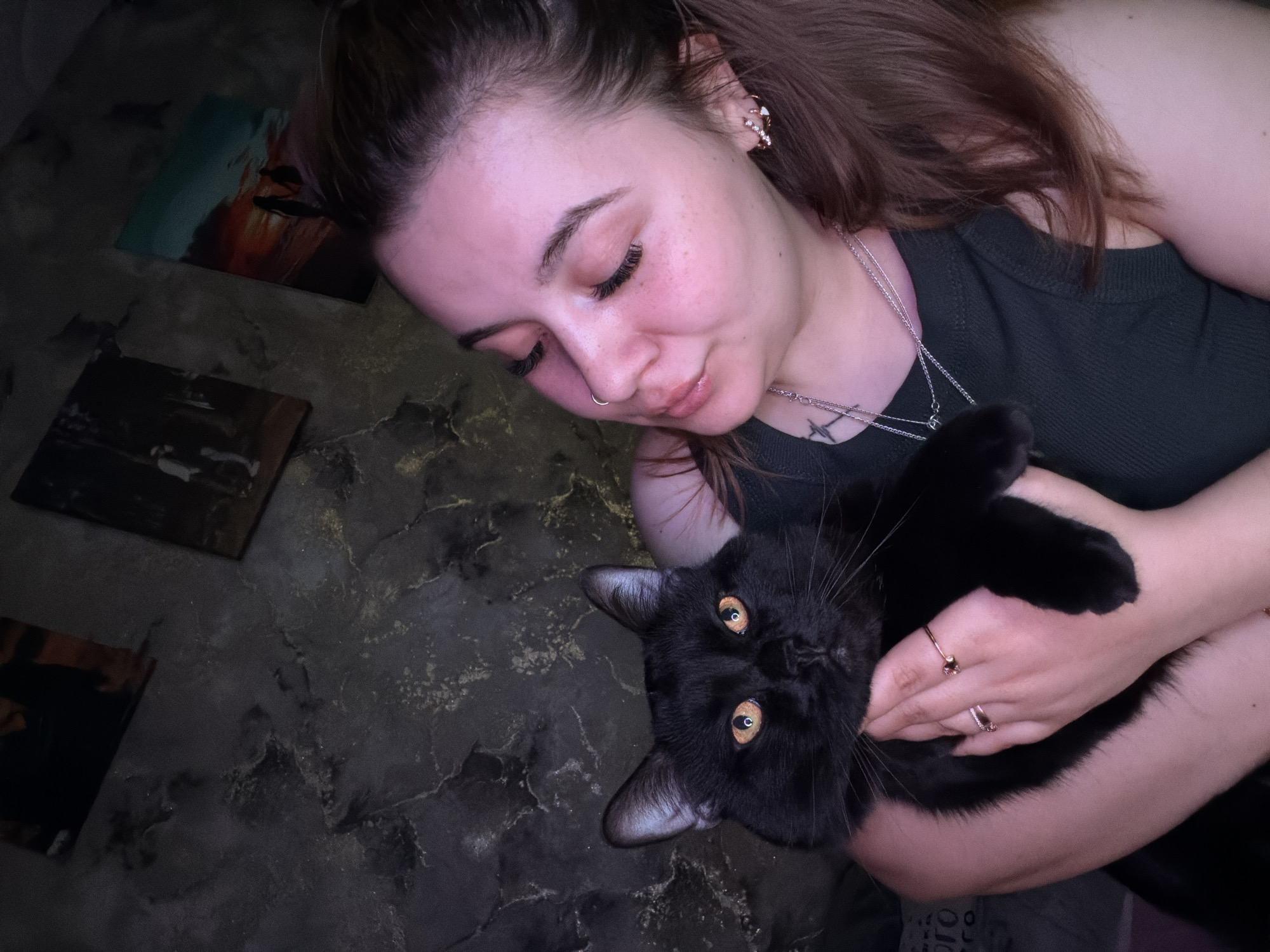 A woman is holding a black cat. She is fair-skinned with brown hair pulled back in a ponytail, a nose ring, and ornate earrings. She wears a dark green shirt and several rings. She has a small tattoo on her chest. The cat has yellow eyes. The background is a gray marble floor. There are two square objects on the floor, possibly picture frames. The location is Chaikovsky, Russia.