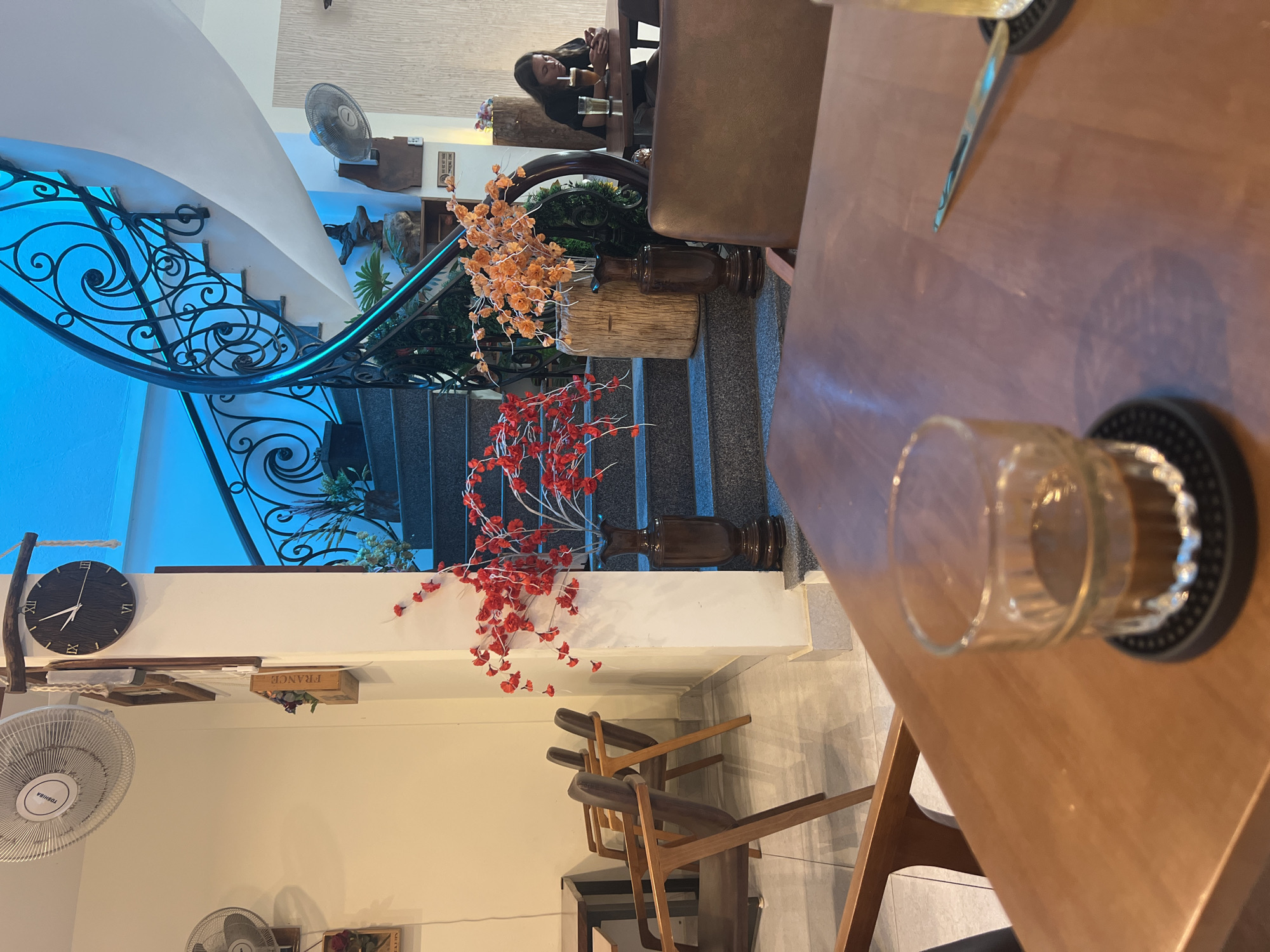 A slightly angled indoor shot focuses on the corner of a wooden table with a glass and a spoon on a black coaster in the foreground. The blurred background features the edge of a staircase with a decorative, wrought-iron railing painted blue on the left side of the frame. Orange and red artificial flowers are situated on top of the steps and landing. The walls are white, and the lower left section shows a wall-mounted fan and a shelf with a clock and decorative box. A person is vaguely visible in the top-left corner of the frame.