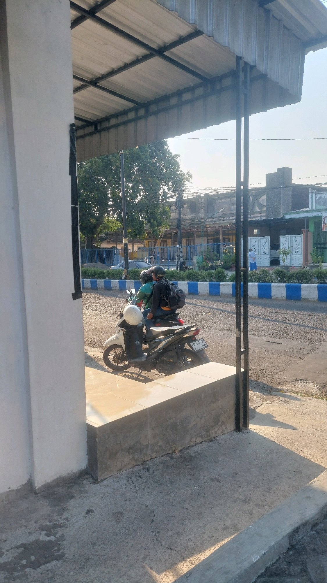 In Nologaten, Indonesia, two individuals are seated on a dark-colored scooter, parked on a concrete ledge. The person in the rear, wearing a black jacket and a dark helmet, has a black backpack secured to the scooter. The individual in front, wearing a green jacket and a light-colored helmet, is also on the scooter. A white helmet rests on the front of the scooter. Behind them, a road is visible with blue and white striped curbing, and beyond that, buildings and trees are present. The scene is illuminated by sunlight, casting shadows on the ground.