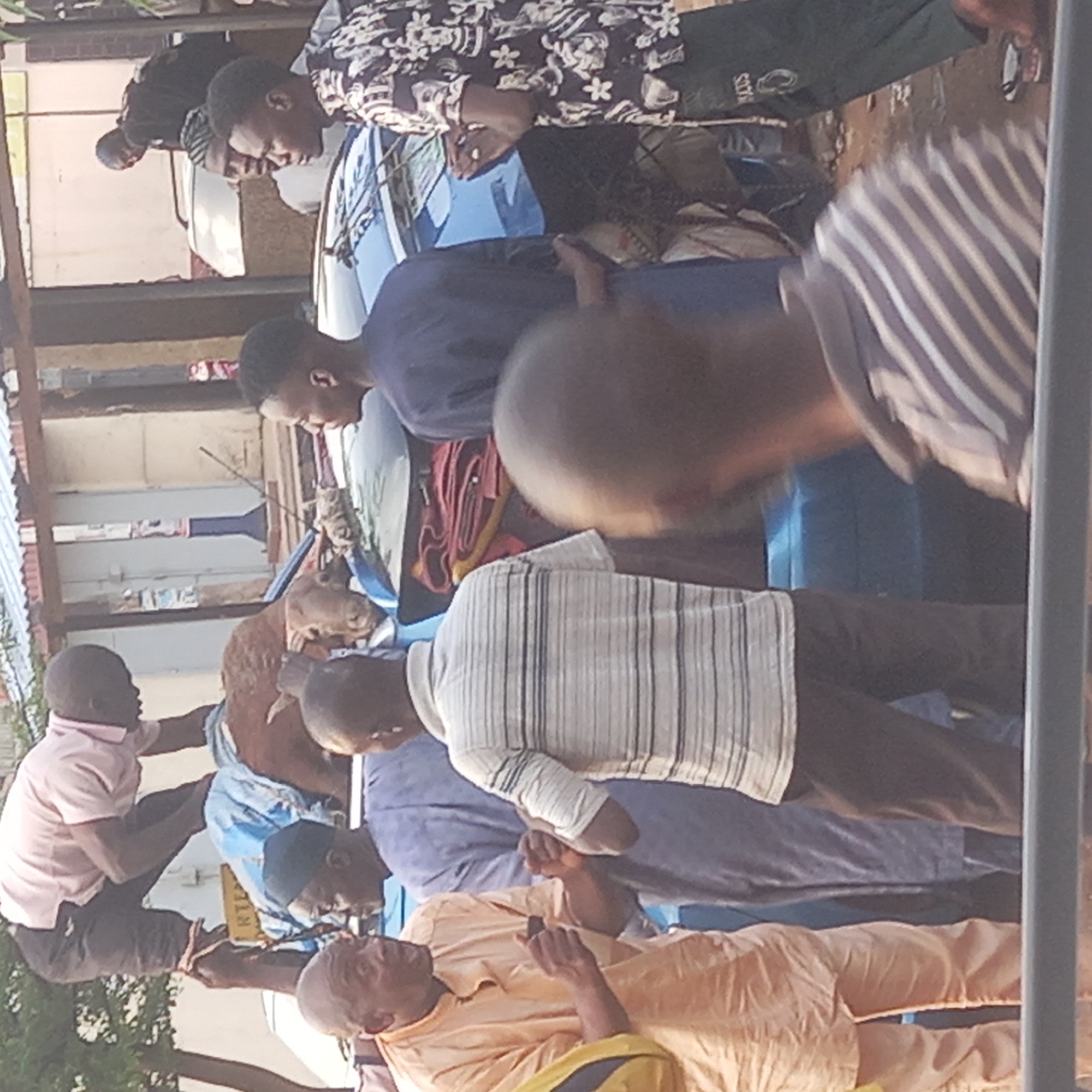 A group of approximately 10 men are gathered around a brown and white goat. The location appears to be outdoors in Dadi Riba, Nigeria. The men are wearing a variety of casual clothing. Some are holding the goat. A blue car is visible behind some of the men. Other visible objects include metal supports, part of a building, and street debris. The men appear to be involved in some sort of interaction involving the goat, but the specific action is not discernible from the image.