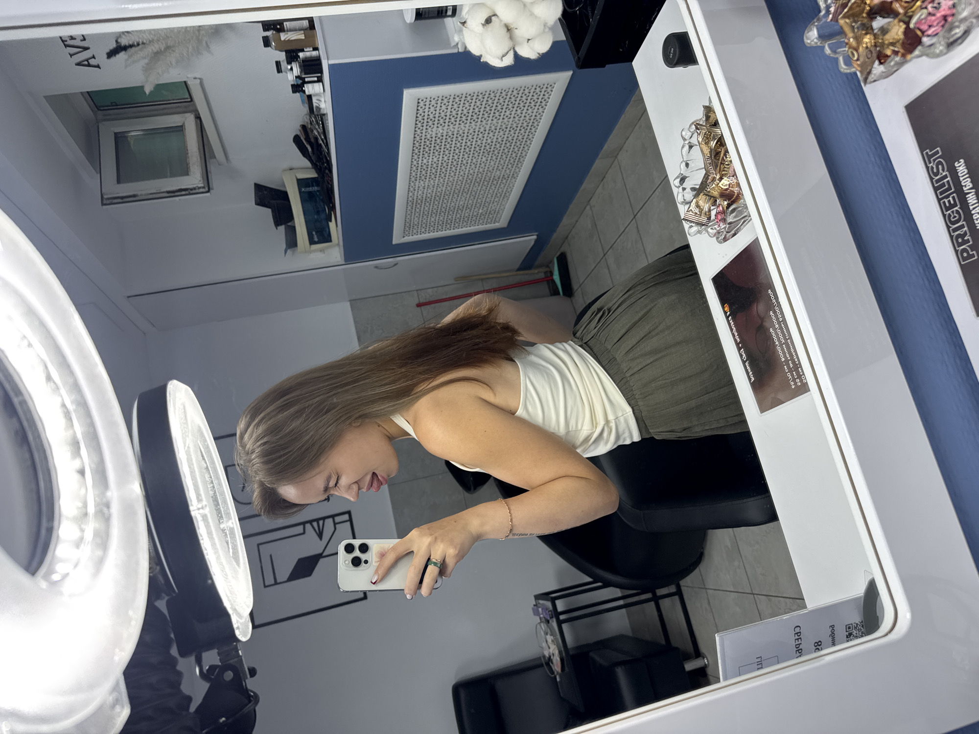 This media file shows a young woman taking a selfie in a salon mirror. She has long, brown hair and is wearing a white tank top and a green pleated skirt. She is holding a white smartphone in her right hand, and her left arm is resting on her lap. A ring light is visible in the foreground, suggesting the photo was taken indoors, likely in a beauty salon, given the presence of various products on shelves in the reflection and a "PRICE LIST" sign for "KERATIN/BOTOKS" services. The setting appears to be a modern salon with blue and white accents on the wall. The woman has a playful expression, with her tongue sticking out slightly. The time of day is not discernible, but the indoor lighting indicates it's not a daytime exterior shot. Text visible in the reflection includes "AVE" and a price list in Russian.