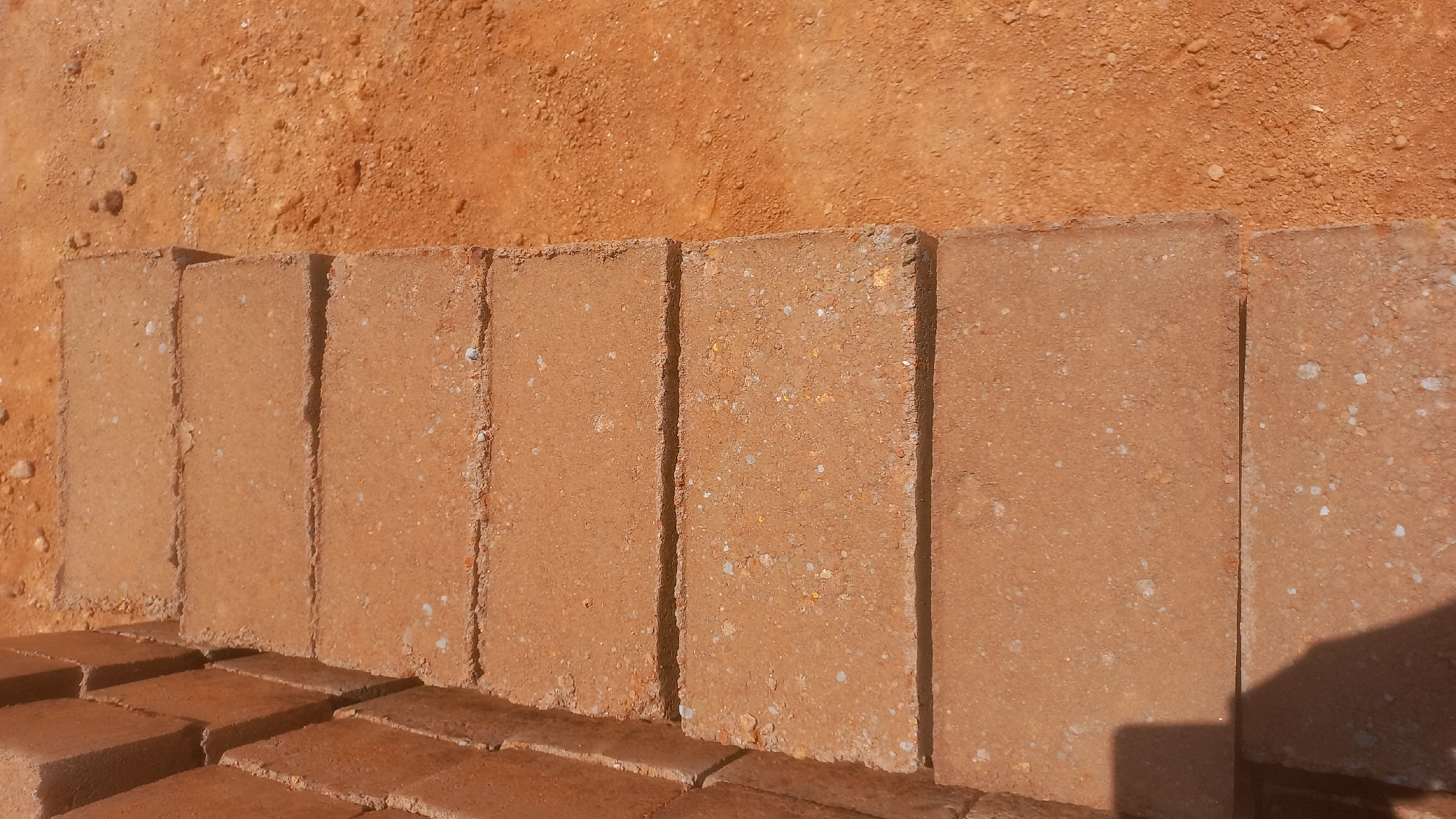 The image shows a close-up, overhead view of a stack of reddish-brown bricks laid out on a dusty, orange-brown ground. The bricks are rectangular and appear to be made of compressed earth or clay, with small white and yellow flecks visible on their surfaces. They are arranged in a row, with their long sides facing the viewer, and are casting shadows. Some of the bricks are stacked higher, forming a more complex arrangement in the foreground. The ground beneath and around the bricks is rough and granular, suggesting an outdoor, possibly construction or storage area. The lighting indicates it is daytime, with the sun casting distinct shadows, suggesting a clear sky. The overall scene is one of construction materials ready for use in Katon Rikkos, Nigeria. There is no visible text in the image.