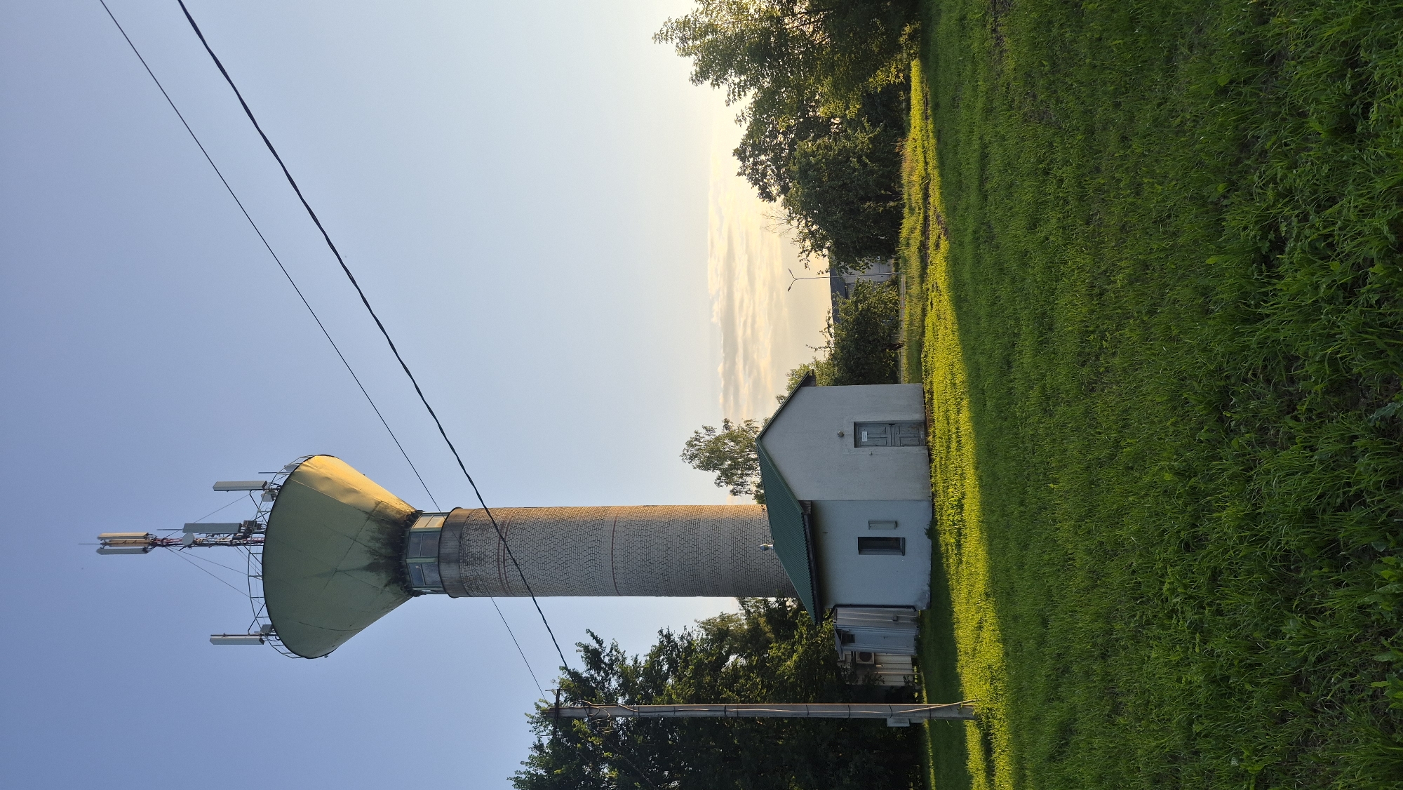 The image shows a water tower with telecommunications antennas on top, located in Krauja, Latvia. A small, white building with a green roof sits at the base of the tower. The scene is illuminated by the warm light of sunset, casting long shadows across the green grass. Trees surround the area, and a power line runs diagonally across the sky. The sky is clear with a few scattered clouds near the horizon. There are no people visible in the image.