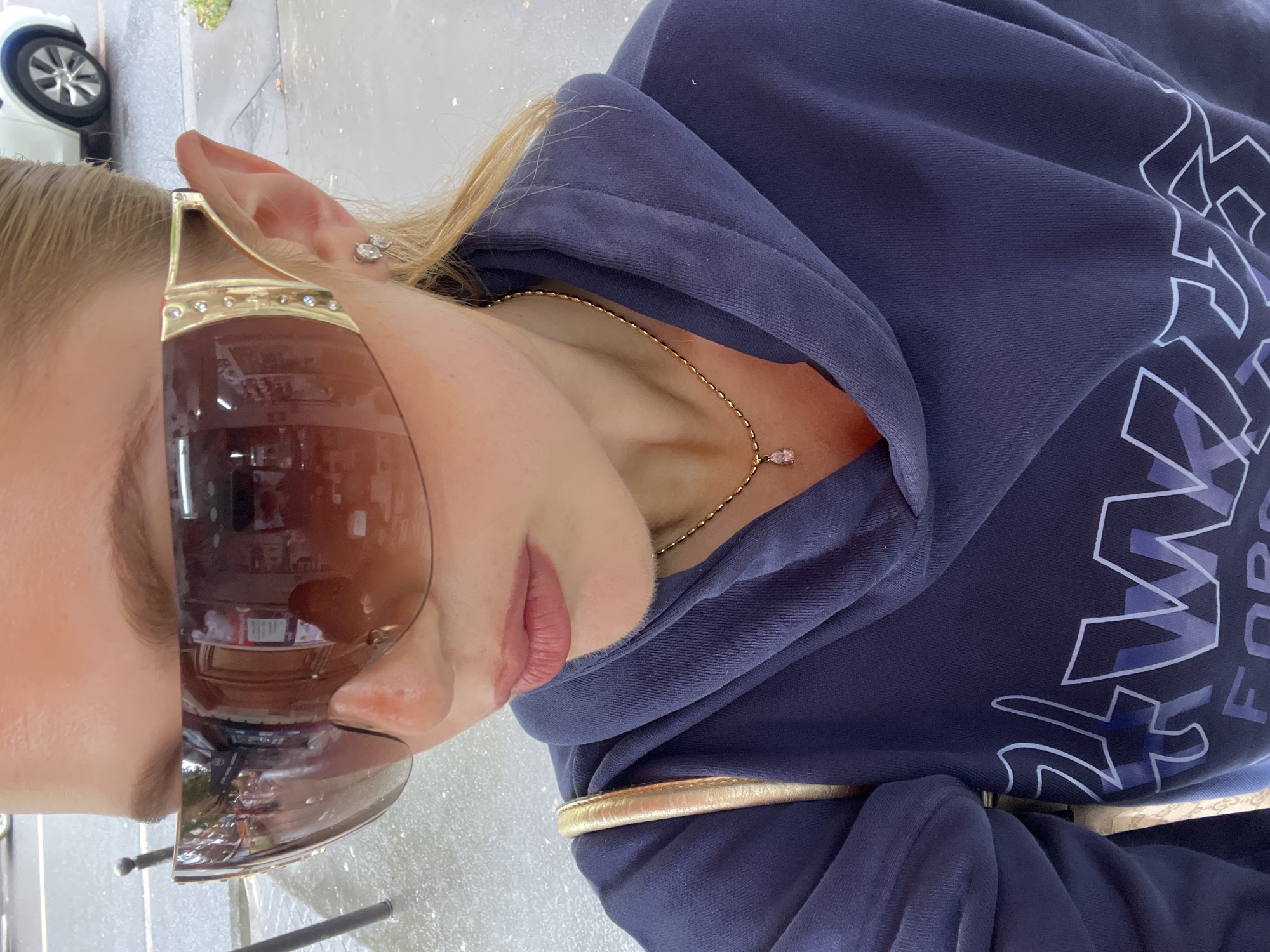 A woman wearing large, gold-framed sunglasses and a blue hooded sweatshirt is positioned outside in a city environment. Her blonde hair is pulled back, revealing an earring. A gold chain necklace with a pendant sits just below the edge of her sweatshirt's hood. The reflected image in her sunglasses shows parts of the surrounding location, including vehicles, people inside of a building, and other reflections suggesting a bustling city scene. Given the reflections of a city and the European plates on the vehicles, it could be plausibly located in a large European city, such as Paris, France.