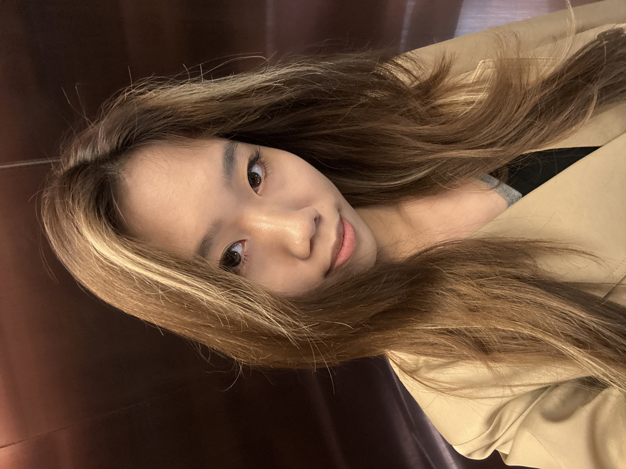 A woman is facing the camera. She has long brown hair with blonde highlights. Her eyes are dark and her complexion is fair. She is wearing a light brown jacket. The background appears to be a dark brown wall.
It is impossible to determine the location of the photograph within the city of West Station Subdistrict, China, without additional information.