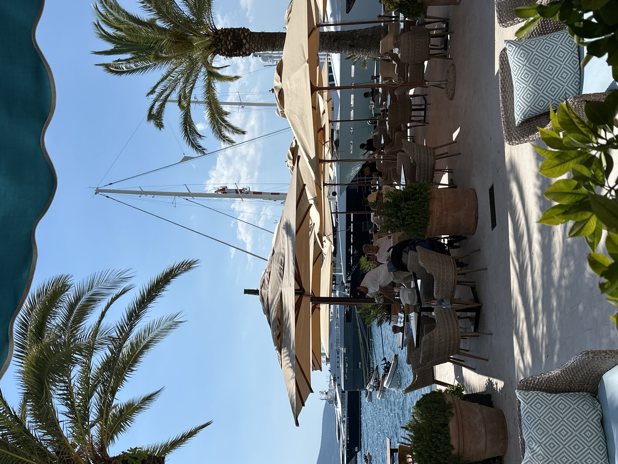 In the city of Donja Lastva, Montenegro, a waterfront restaurant scene unfolds under a bright, sunny sky with scattered clouds. Large, tan umbrellas provide shade over several outdoor dining tables, many of which are occupied by patrons. Waitstaff, identifiable by their attire, attend to the tables. In the background, the blue water of the bay is dotted with various boats, including sleek yachts docked along the waterfront. Palm trees with lush green fronds frame the scene, adding to the tropical, relaxed atmosphere. The pavement is light-colored and appears clean, with shadows cast by the umbrellas and foliage. The overall ambiance is one of leisure and enjoyment in a picturesque coastal setting.