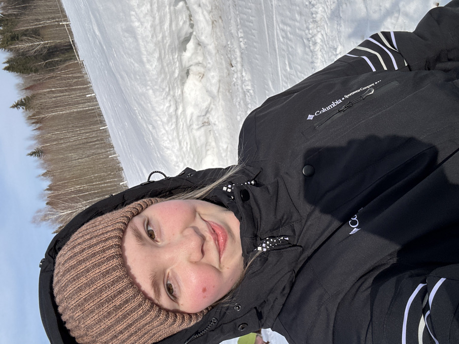 An individual, identified as a female, is depicted in a close-up outdoor photograph taken in Lesnoy, Russia. She wears a light brown ribbed knit beanie and a black hooded winter jacket, featuring a "Columbia Sportswear Company" logo on the right chest and three white horizontal stripes on the left sleeve. Her blonde hair is partially visible beneath the beanie. She has a faint smile and rosy cheeks, with a small mole present on her left cheek. The background reveals a snow-covered landscape, sloping upwards towards a forest line composed of both deciduous and coniferous trees under a clear blue sky. The scene is brightly illuminated by natural sunlight.