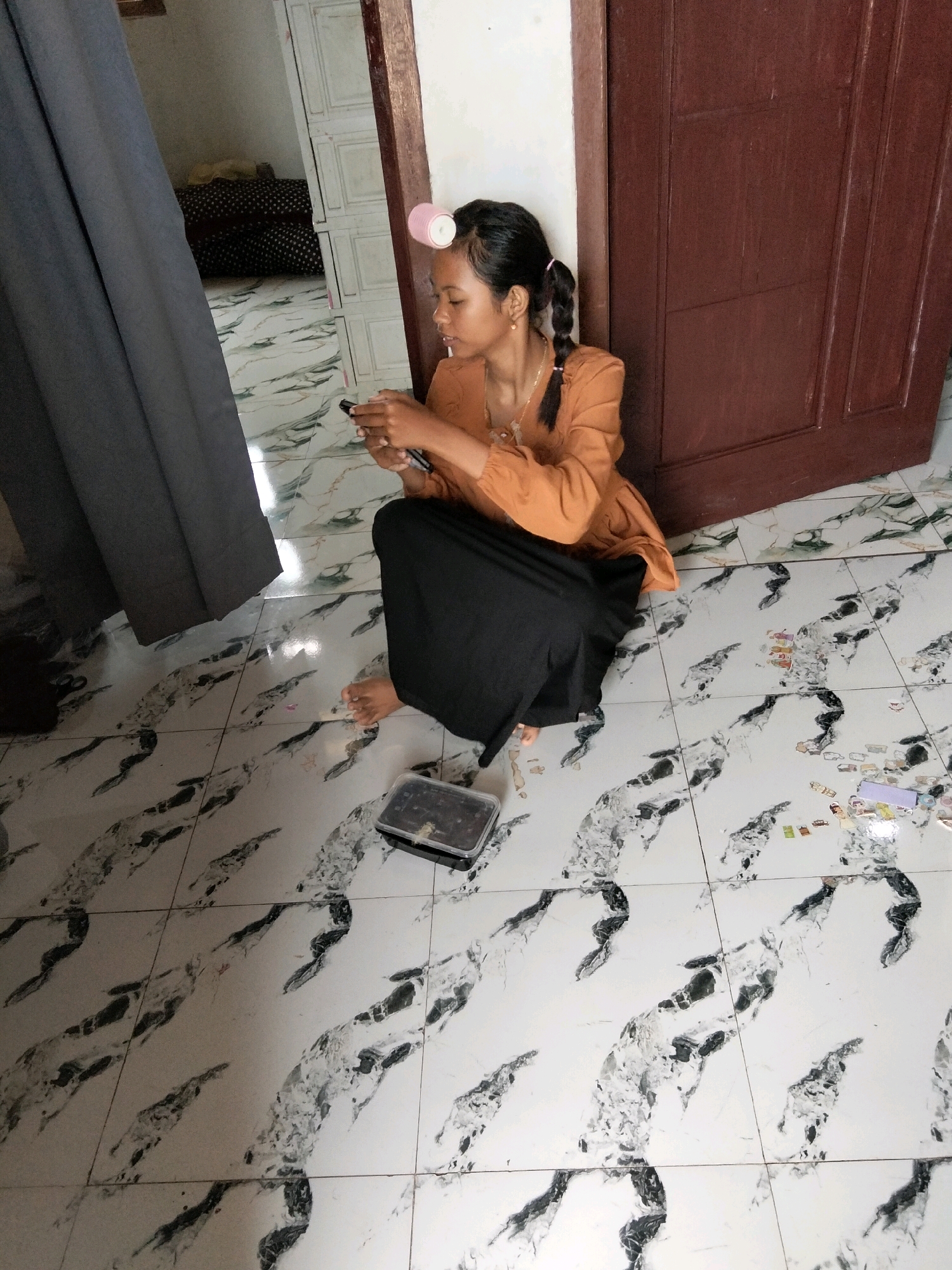A young woman with hair curlers sits on a white tiled floor with black and grey marble-like patterns. She is wearing an orange long-sleeved shirt and a black skirt. Her legs are crossed, and she is holding a mobile phone in her hands, looking down at it. To her right is a dark brown wooden door. In front of her is a small black plastic container. To her left, a grey curtain hangs. In the background, a room with a bed and cabinets is visible. The scene appears to be an interior setting, likely a residential area in Perambahan, Indonesia.
