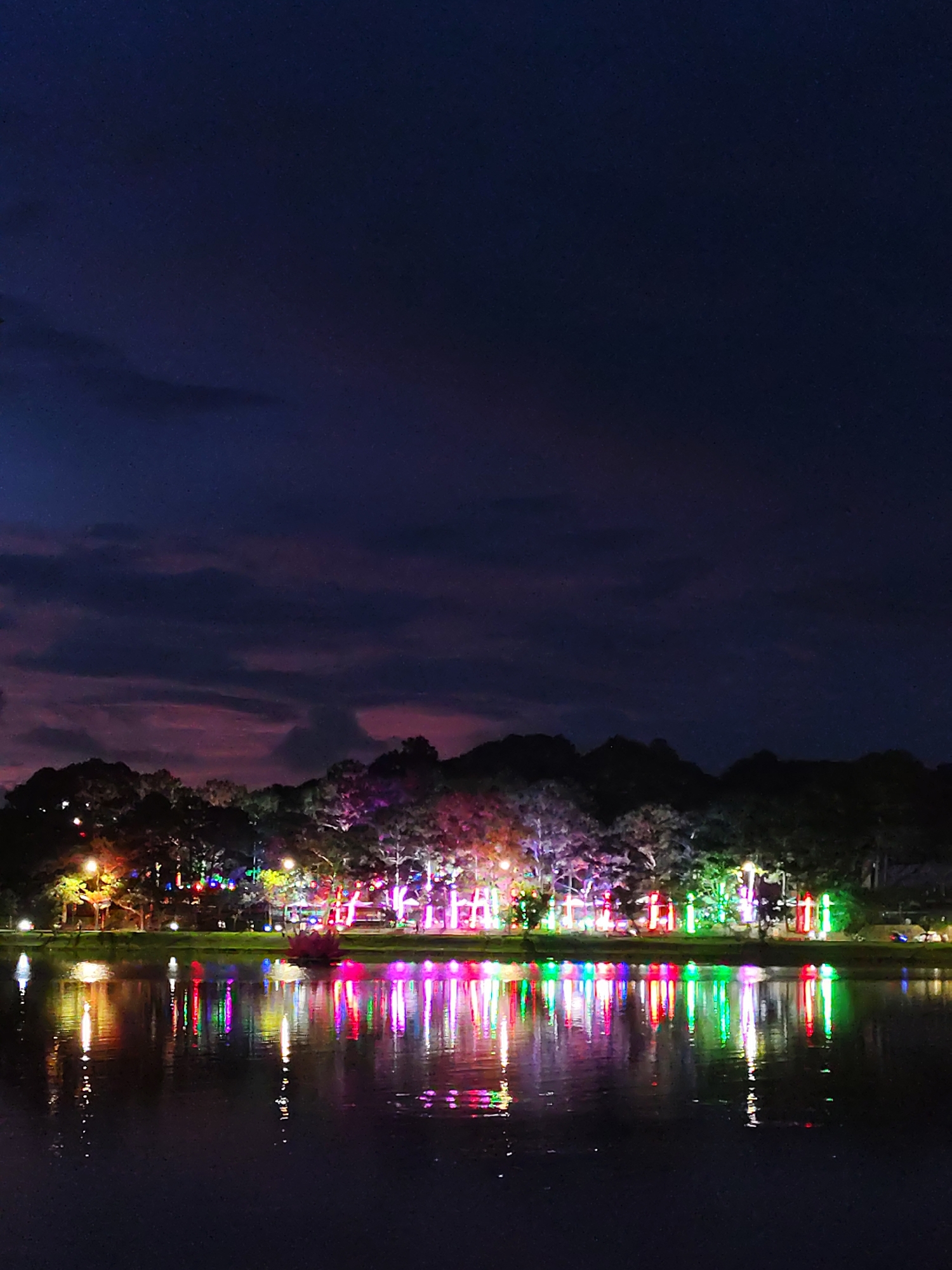 At night, the image depicts a lake in Da Lat, Vietnam. The water surface reflects a series of colorful lights adorning trees along the shoreline. Above the trees, a dark, cloudy sky fades into a deep blue night.