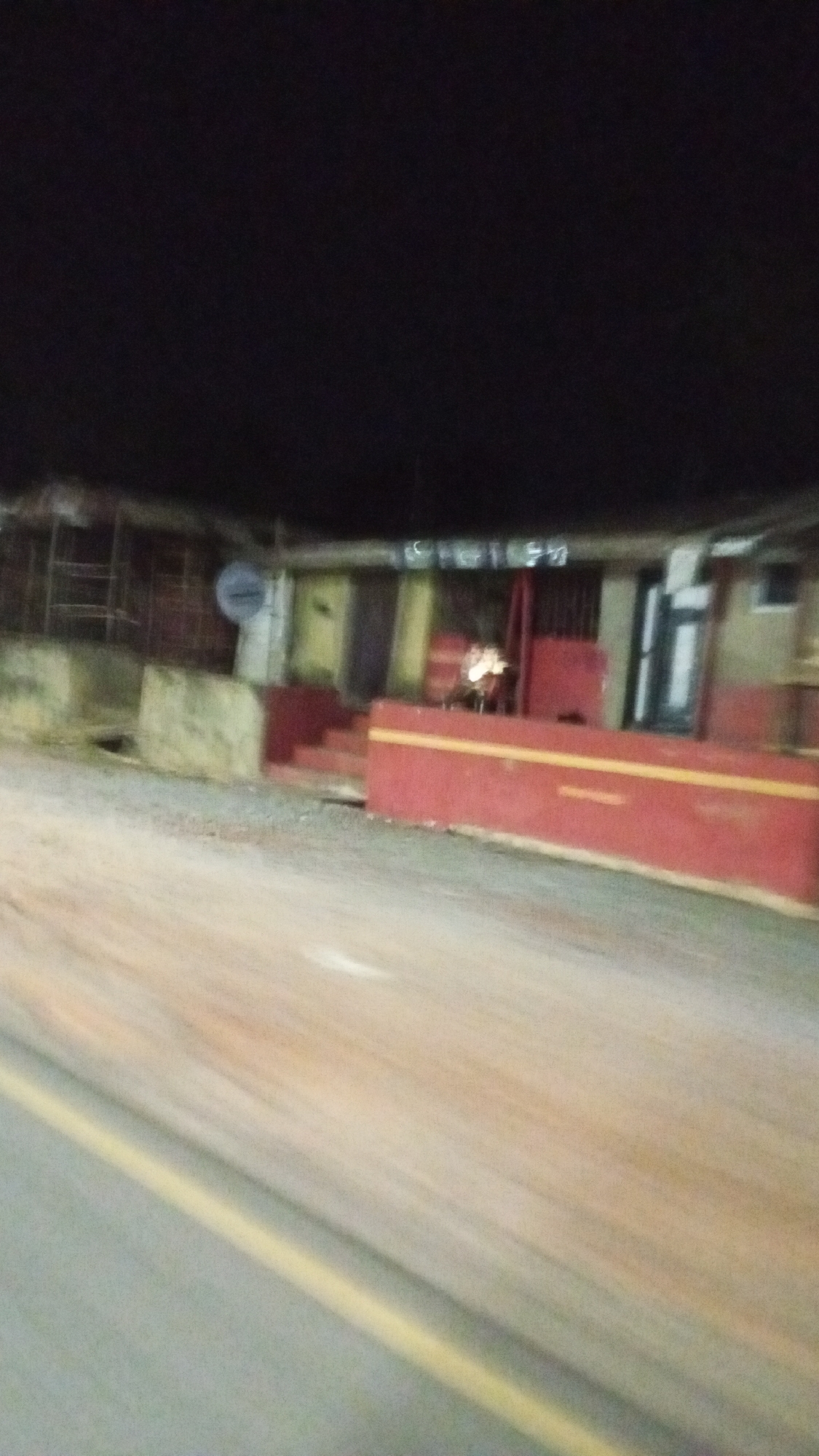 The image shows a street scene at night in Adankolo, Nigeria. A building with a red wall and yellow stripe sits on the side of the road. The building has several doors and windows. A satellite dish is visible on the roof. The road is blurred, likely due to movement during the photo capture. There are no people visible in the image.