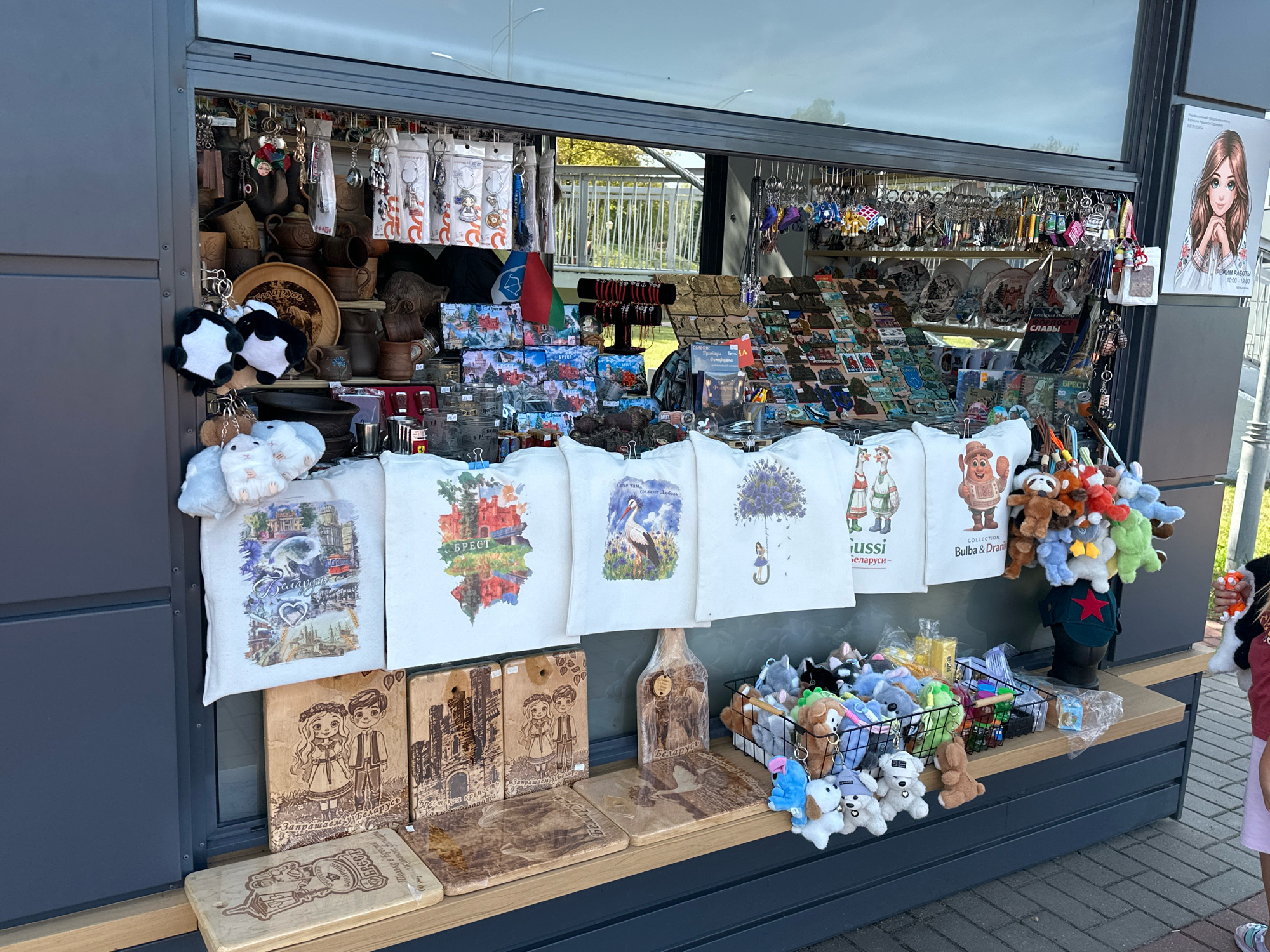 A street kiosk in Brest, Belarus, displays various souvenirs for sale. The kiosk features shelves stocked with trinkets, keychains, plates, and bags. A prominent sign on the right side of the kiosk advertises operating hours from 10:00 to 19:00, with a stylized illustration of a young woman. In the foreground, a collection of plush toys, including stuffed animals and cartoon characters, are arranged on shelves and in baskets. Wooden decorative items, such as cutting boards with carved designs, are also part of the merchandise. The overall scene depicts a vibrant marketplace offering locally themed souvenirs.