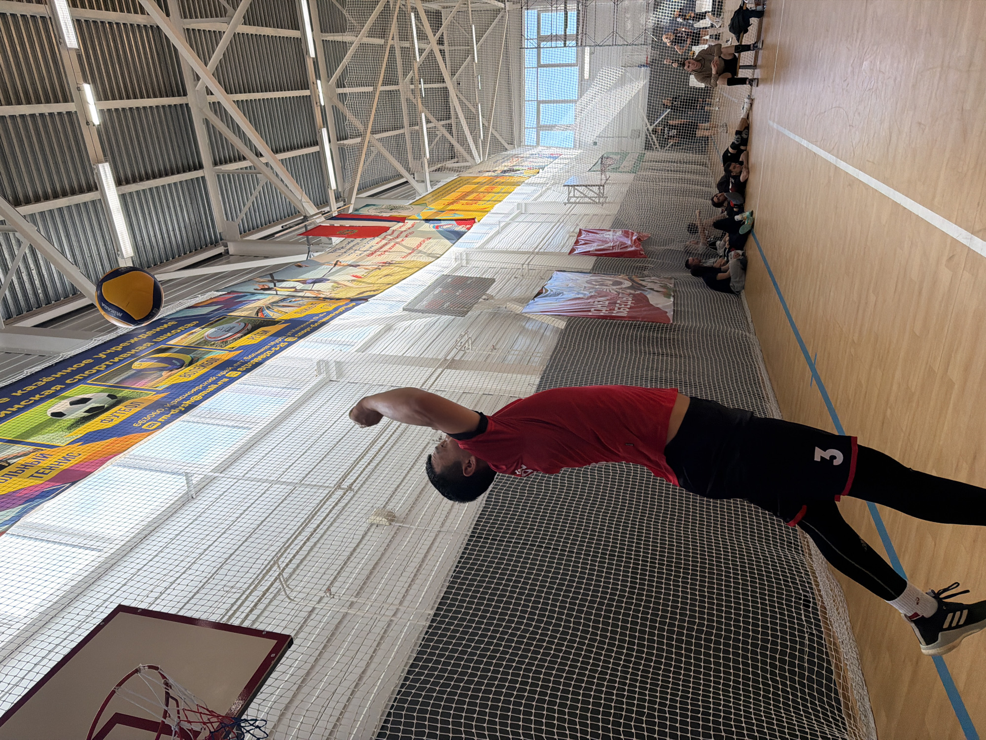 A sports complex in the city of Bolshaya Murta, Russia, is depicted with a man playing volleyball. The man is wearing a red shirt with the number "3" on the back, black shorts, and black leggings. He is arching his back to hit a yellow and blue Mikasa volleyball, which is airborne above a basketball hoop. Banners with sports-related images adorn the walls, including ones with a soccer ball and the word "FOOTBALL" in Russian. A scoreboard is visible overhead, and spectators are seated along the side of the court. The building has a high, metal-framed ceiling covered with netting.