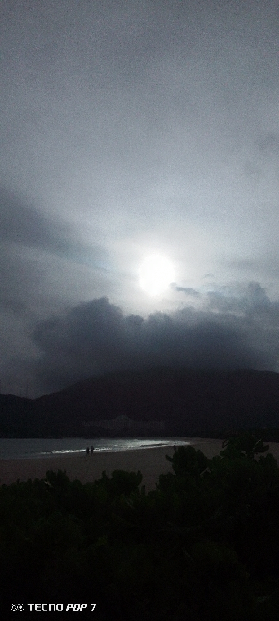 The image shows a beach scene in Pedro González, Venezuela on a cloudy day. The sun is visible through the clouds, casting a soft light on the water. A dark silhouette of a mountain occupies the center of the image. On the beach, there are two people. The foreground contains vegetation. The camera information "TECNO POP 7" is in the bottom left corner.